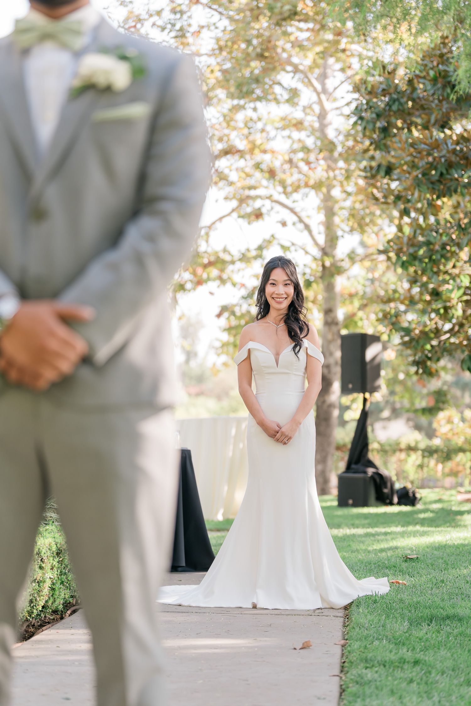 First look wedding photo with bride in white gown standing behind groom in light gray suit outdoors.