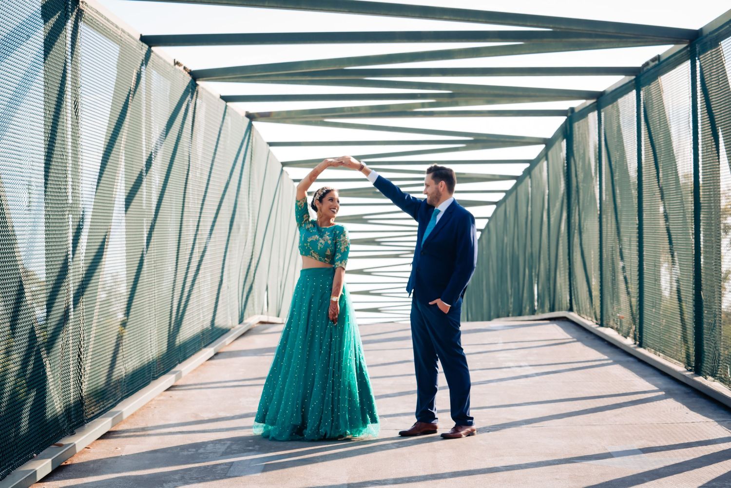 A couple walks through a geometric metal bridge walkway with repeating angular patterns and green accents.