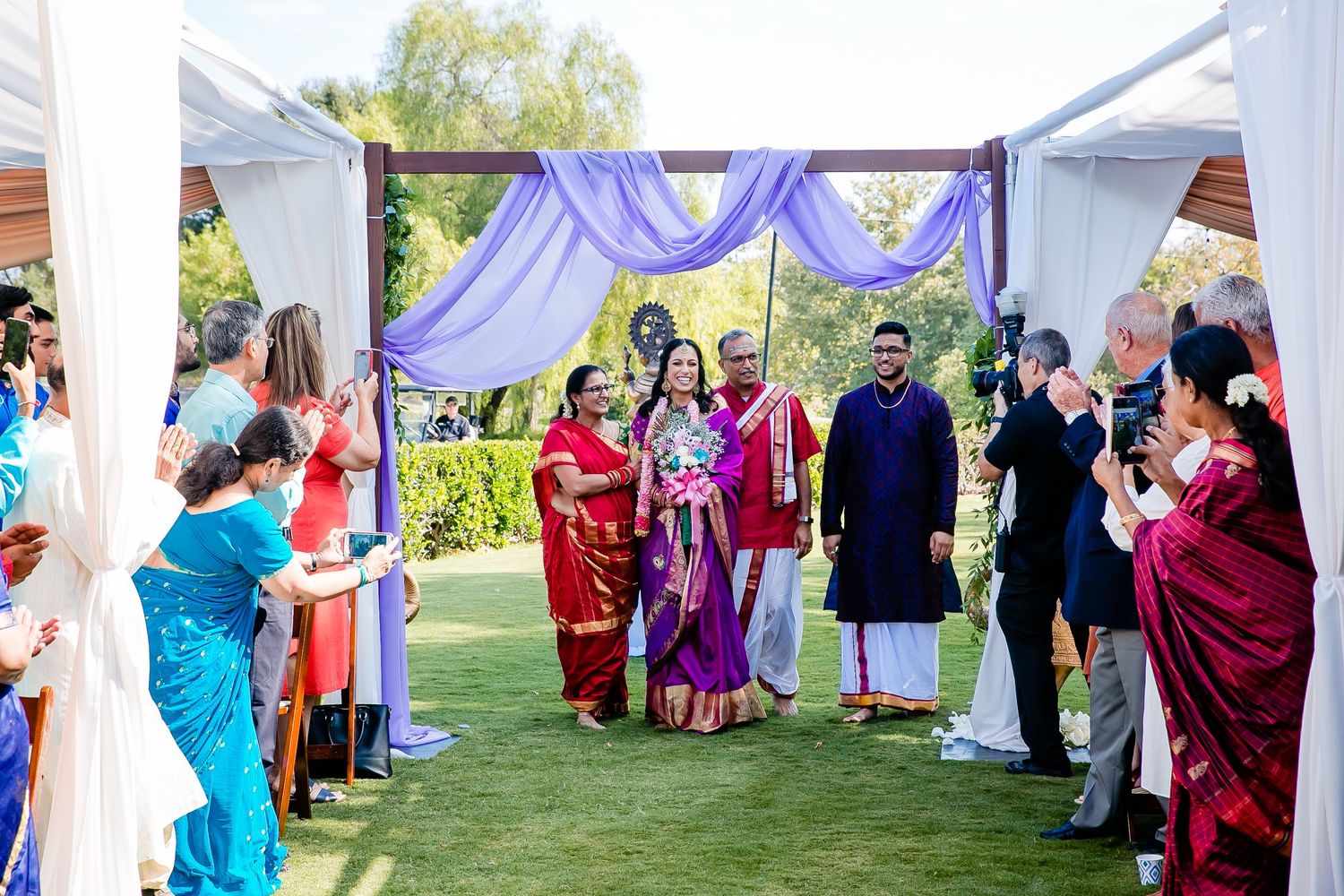 Outdoor wedding ceremony under white tent with guests in colorful traditional ethnic attire standing in rows.