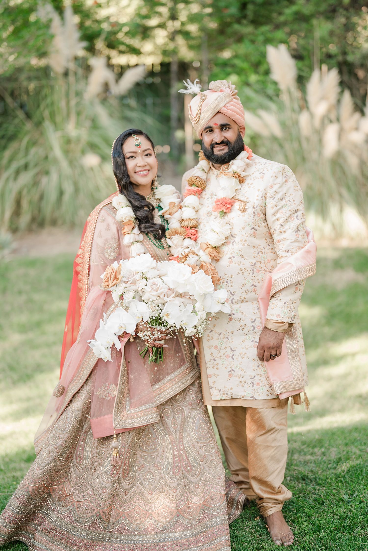 Wedding group in elegant peach and cream traditional outfits pose on grass under trees.