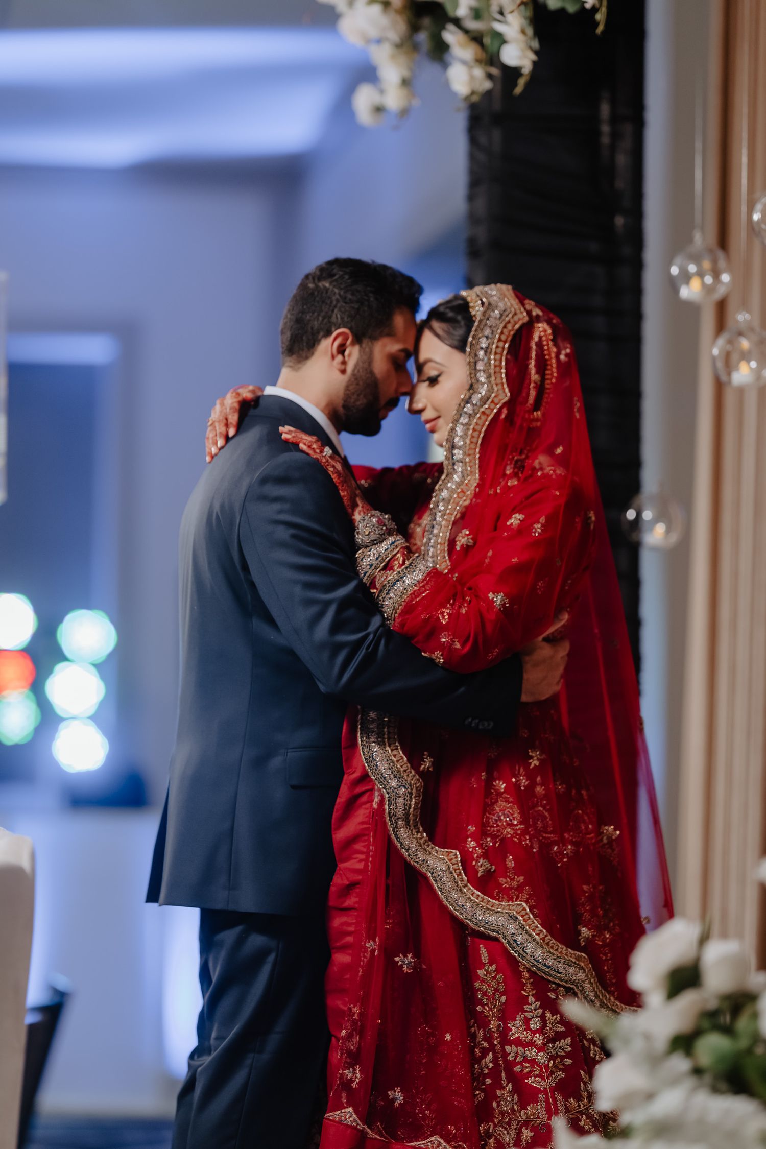 A couple shares an intimate moment at their wedding, with the bride wearing a stunning red and gold traditional bridal lehenga.