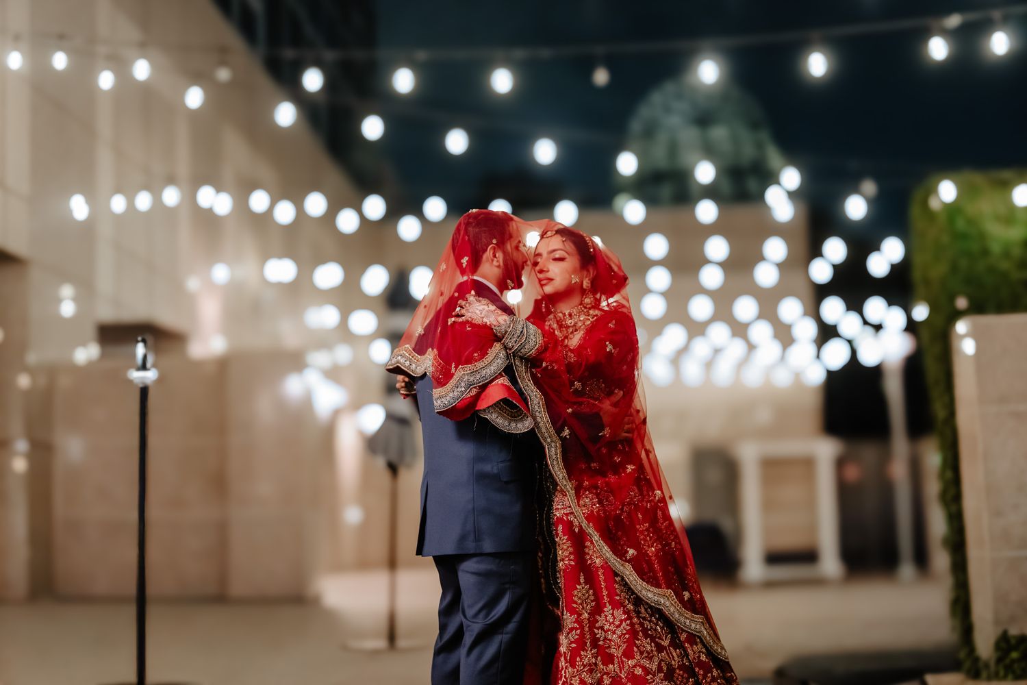 A couple embraces under twinkling string lights at a wedding reception, with the bride wearing a traditional red dress.