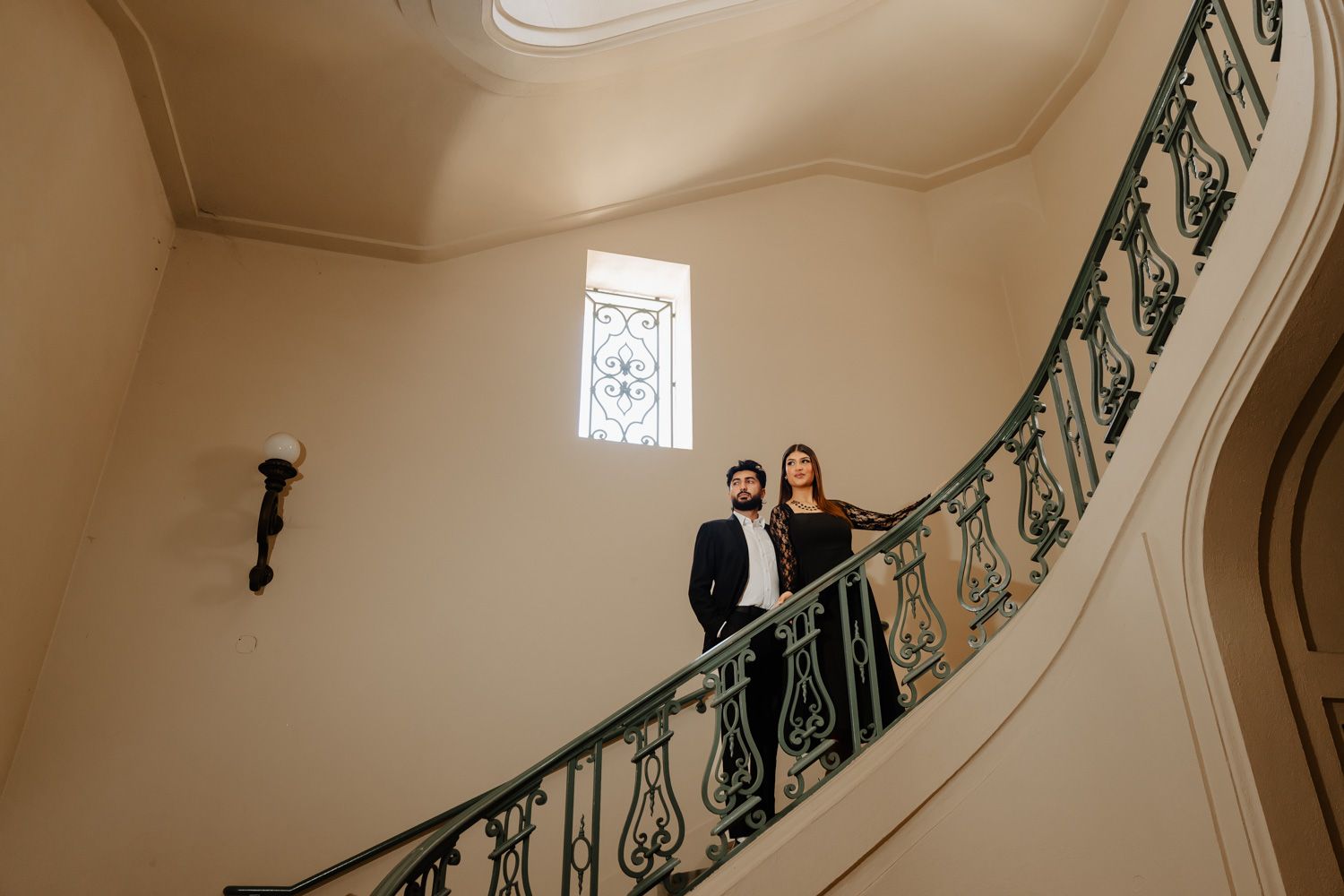 Couple posing on elegant curved staircase with ornate iron railing in a classic interior.