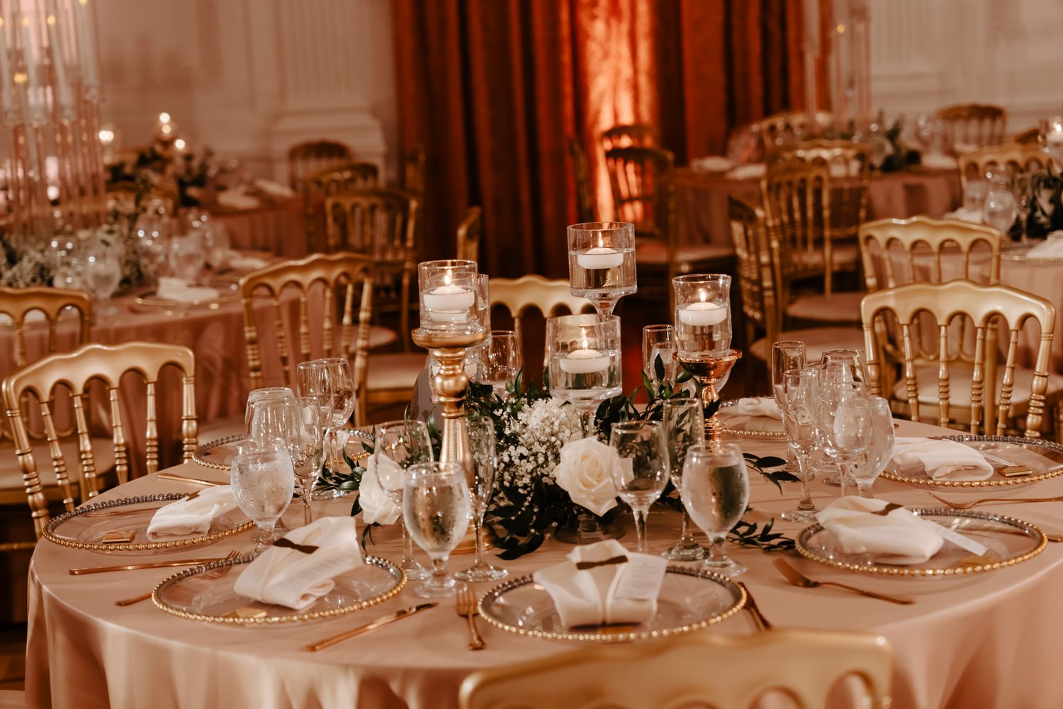Elegant wedding reception tables set with fine china, glassware and golden candelabras in warm lighting.