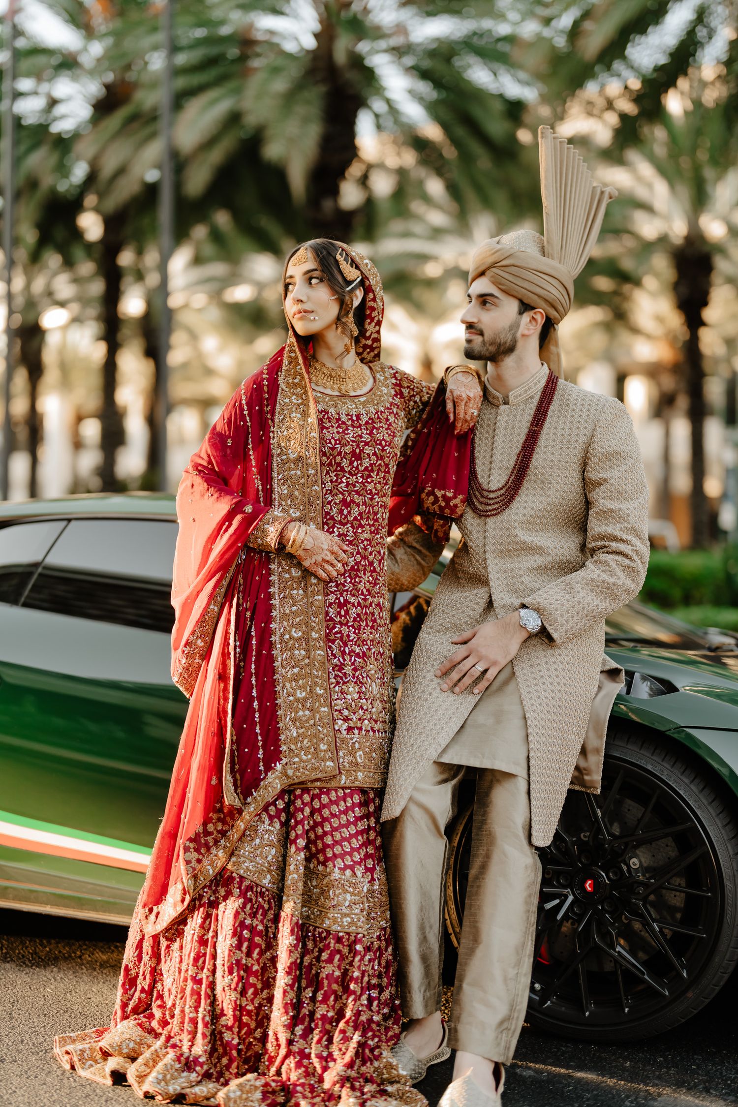 A couple in traditional South Asian wedding attire pose together in front of palm trees and a luxury car.