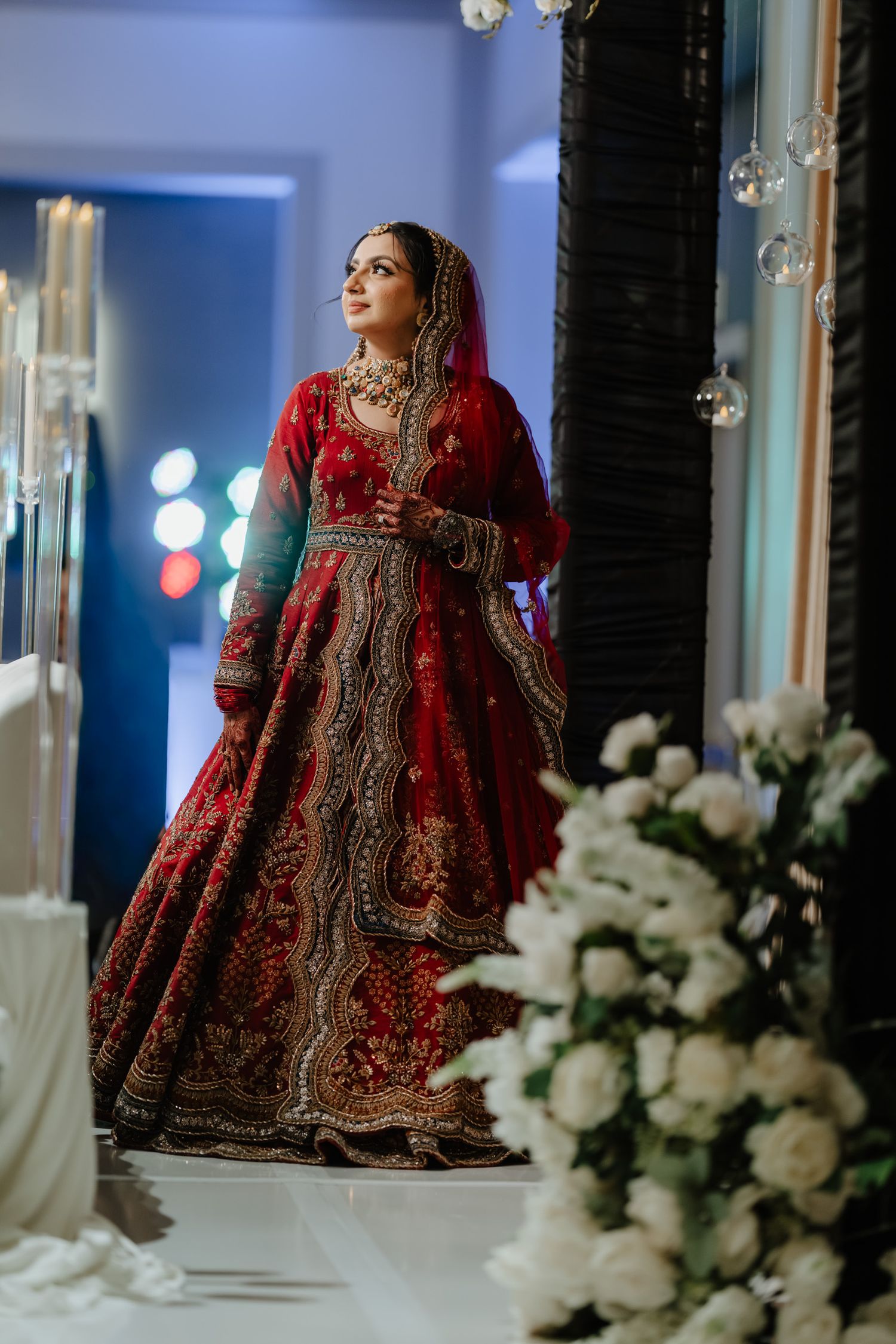 Bride in red and gold embroidered lehenga poses elegantly in a blue-lit room with white floral decor.