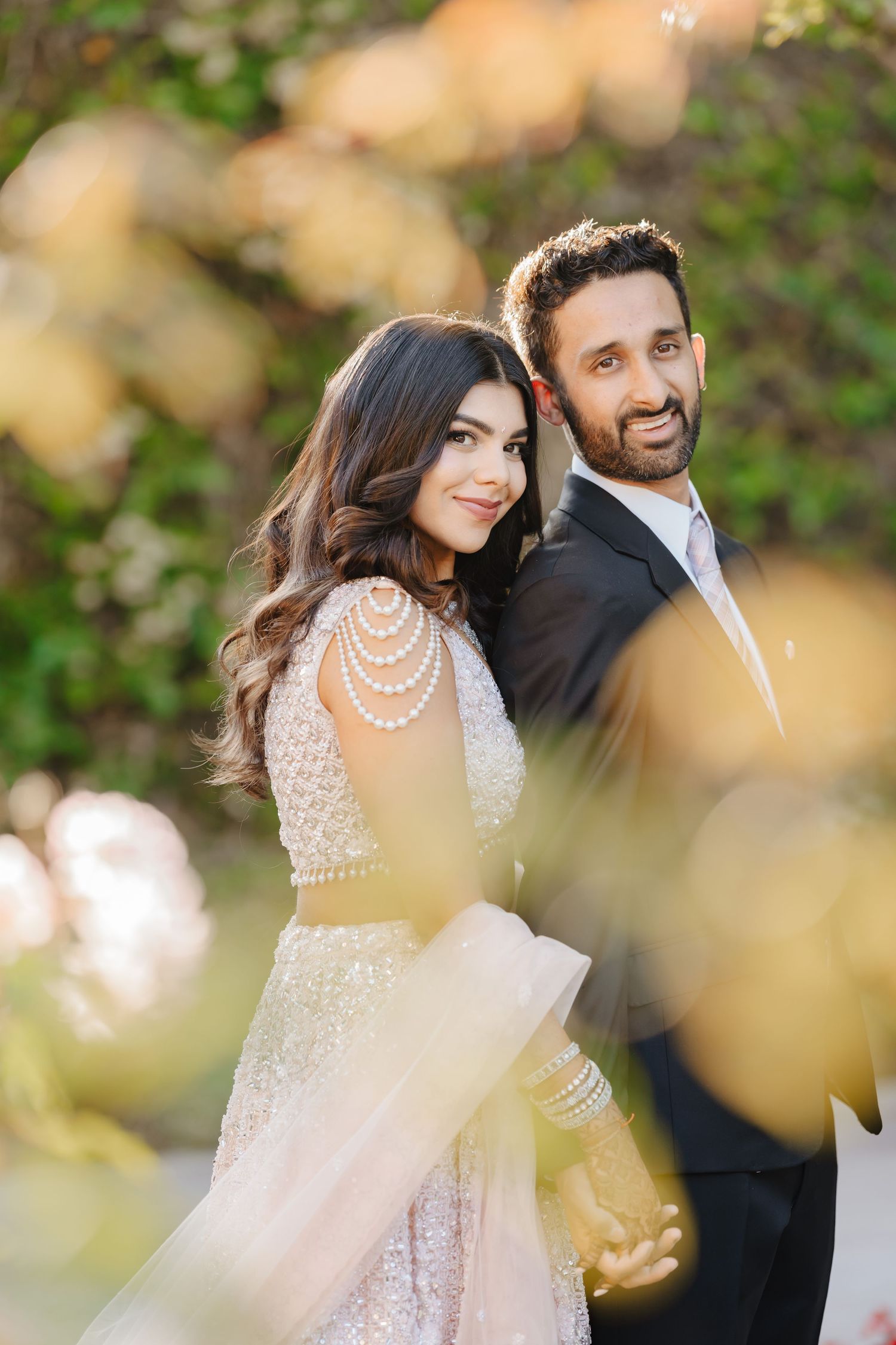 A bride in a beaded gown with pearl necklaces and groom in a suit pose together outdoors at golden hour.