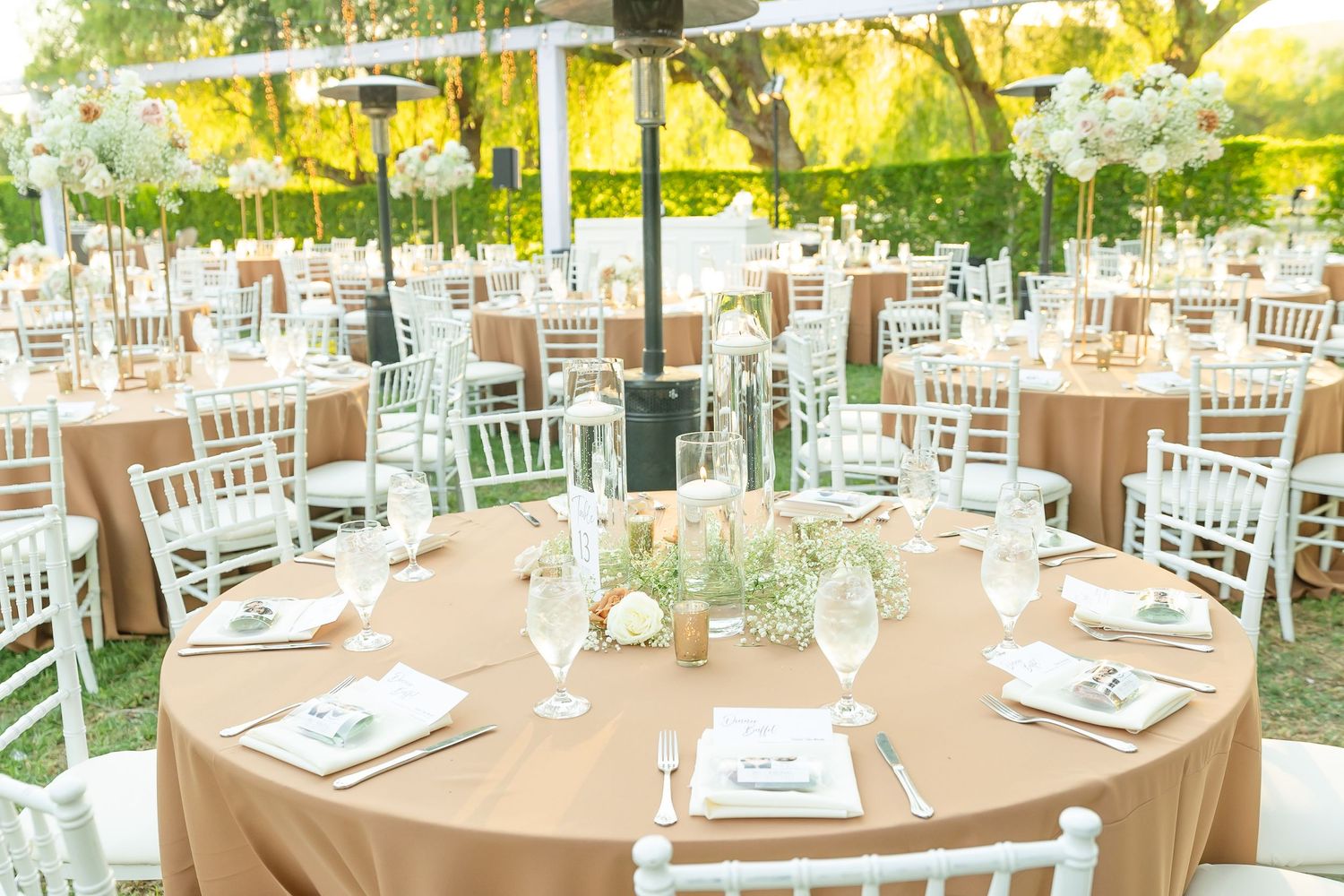 Elegant outdoor wedding reception setup with white chiavari chairs and round tables under natural tree canopy.