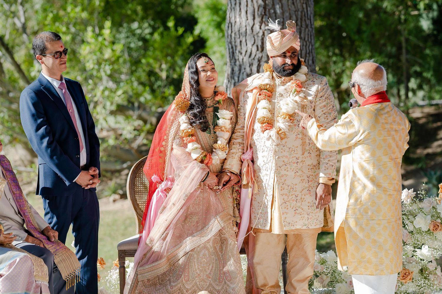 Wedding party members gather outdoors under trees for formal group photos in traditional South Asian attire.
