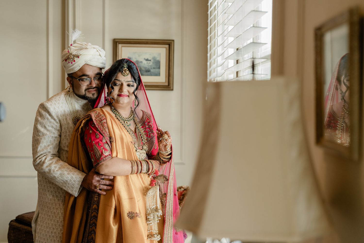 A couple in traditional South Asian wedding attire share an intimate moment by a sunlit window.