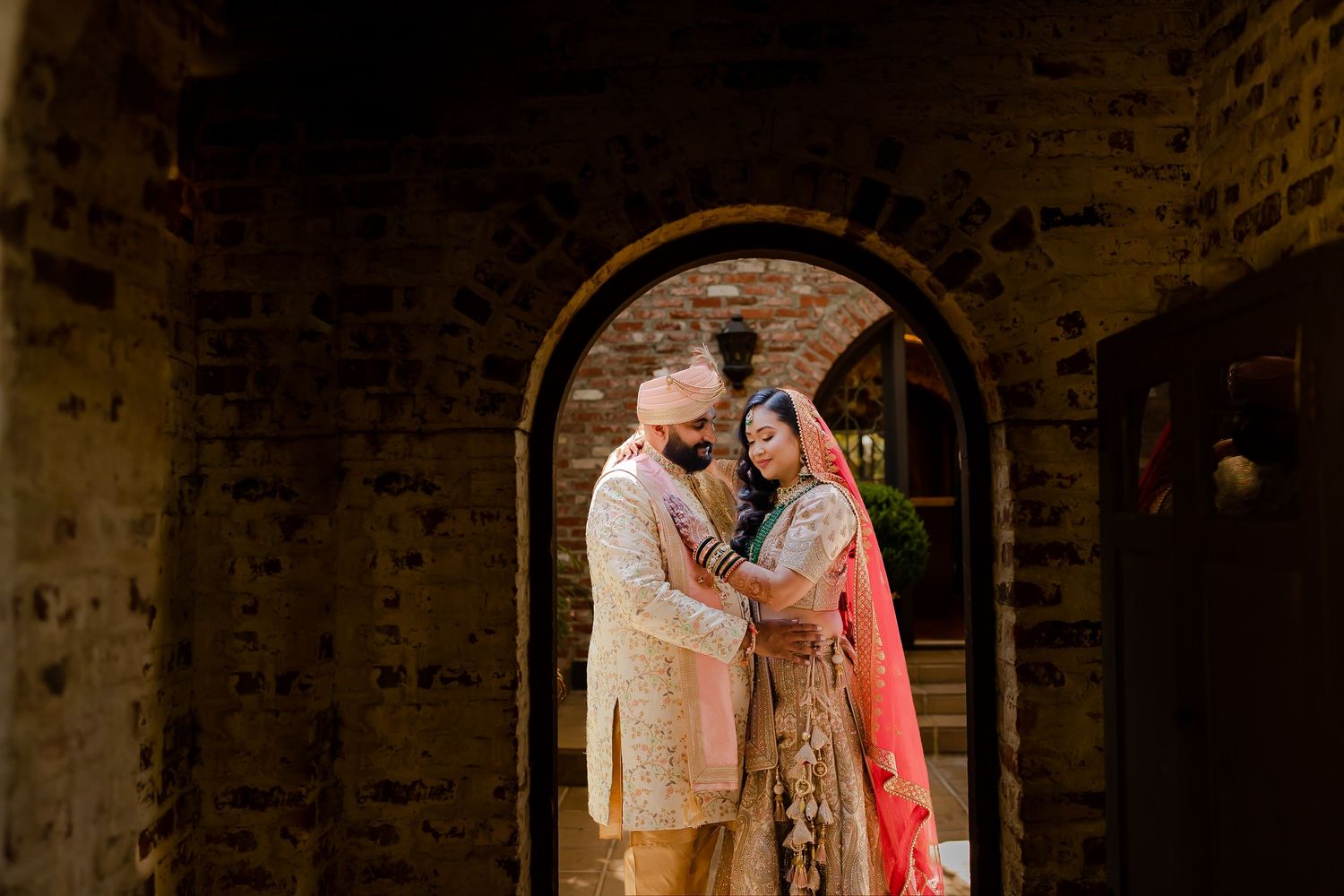 A couple in traditional Indian wedding attire share an intimate moment in a dimly lit rustic setting.