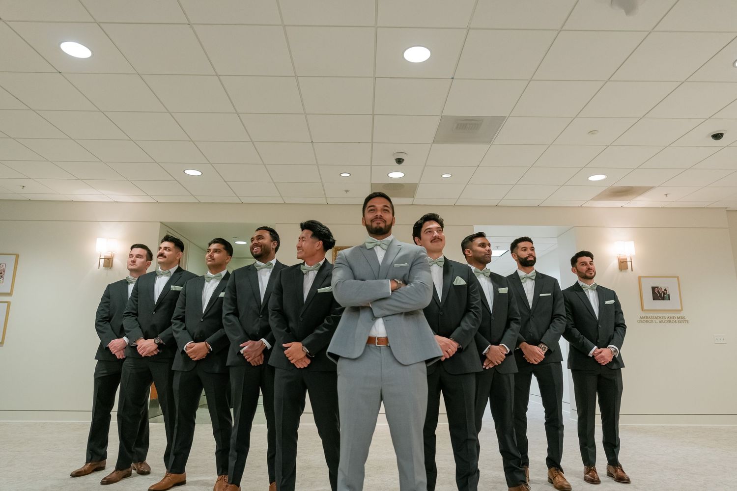 Wedding party photo of groomsmen in black suits with groom in light gray suit standing in elegant indoor venue.