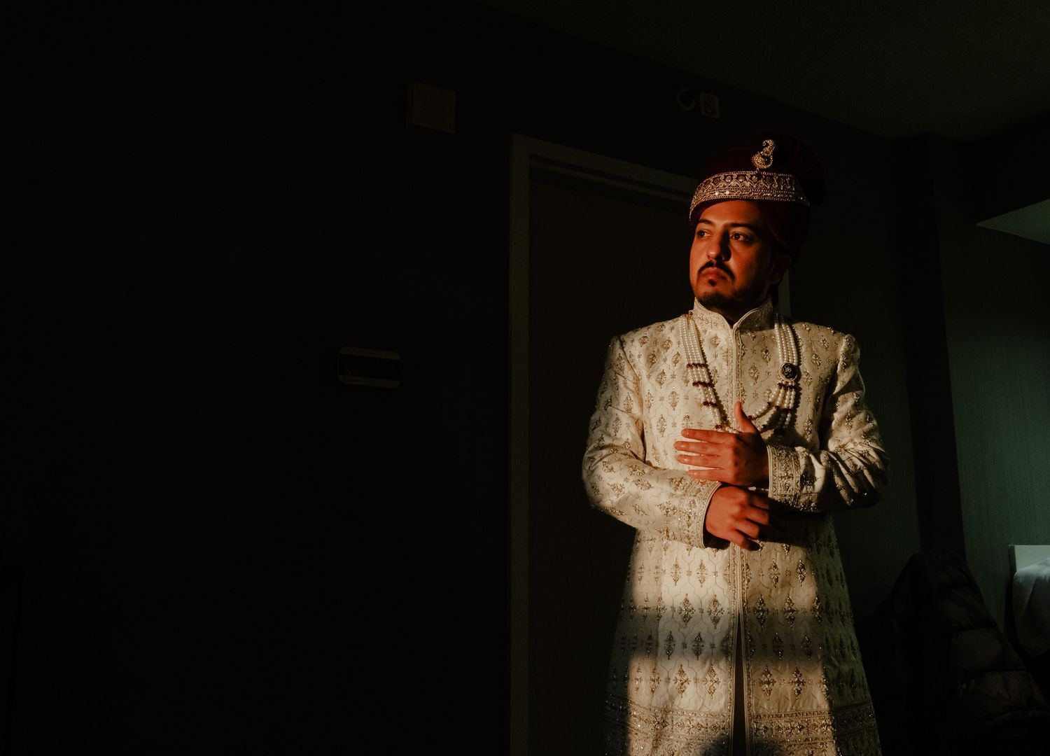 Traditional Indian groom wearing an ornate white sherwani suit in dramatic lighting.