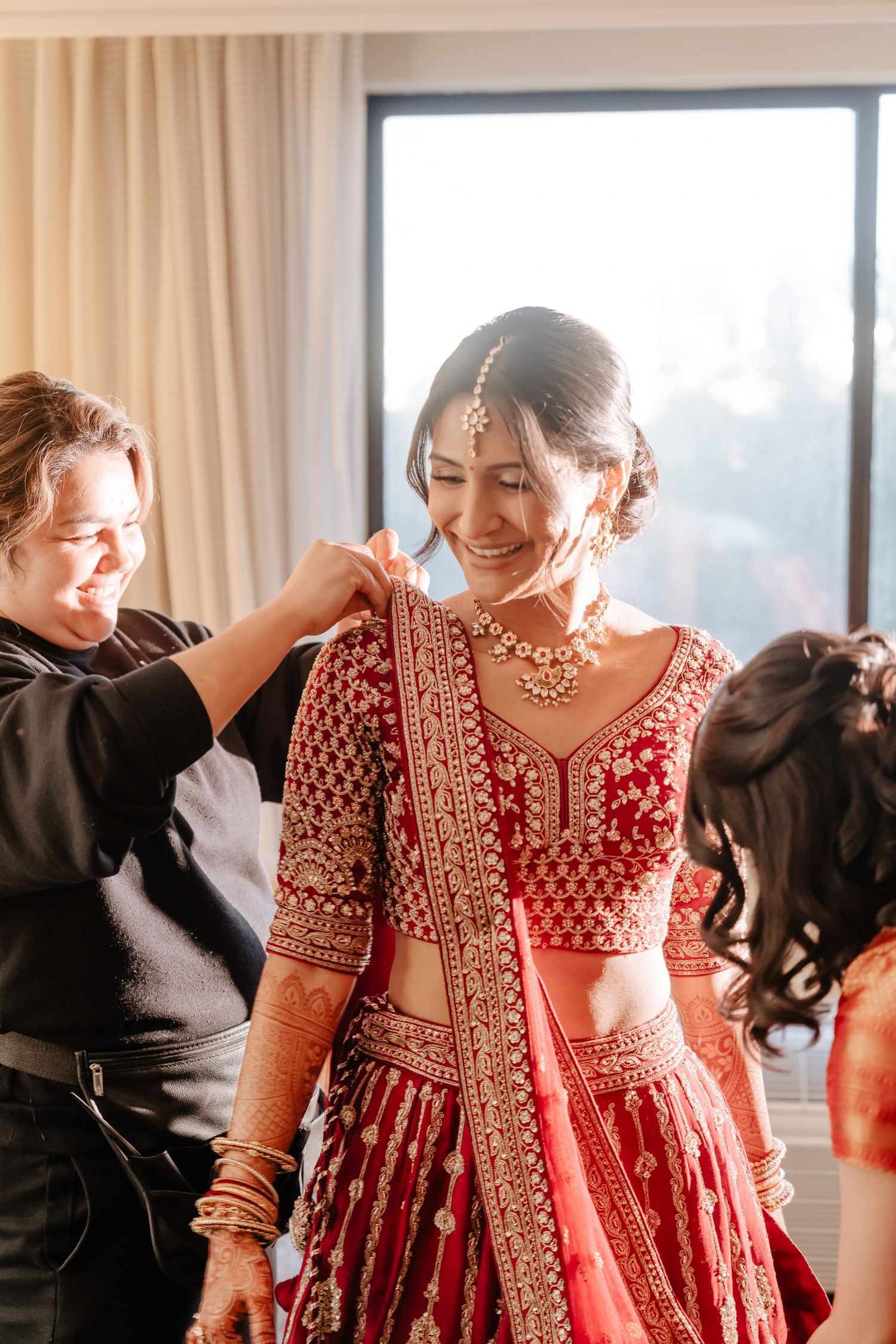 A bride wearing a traditional red and gold embroidered lehenga gets help with final adjustments during wedding preparations.