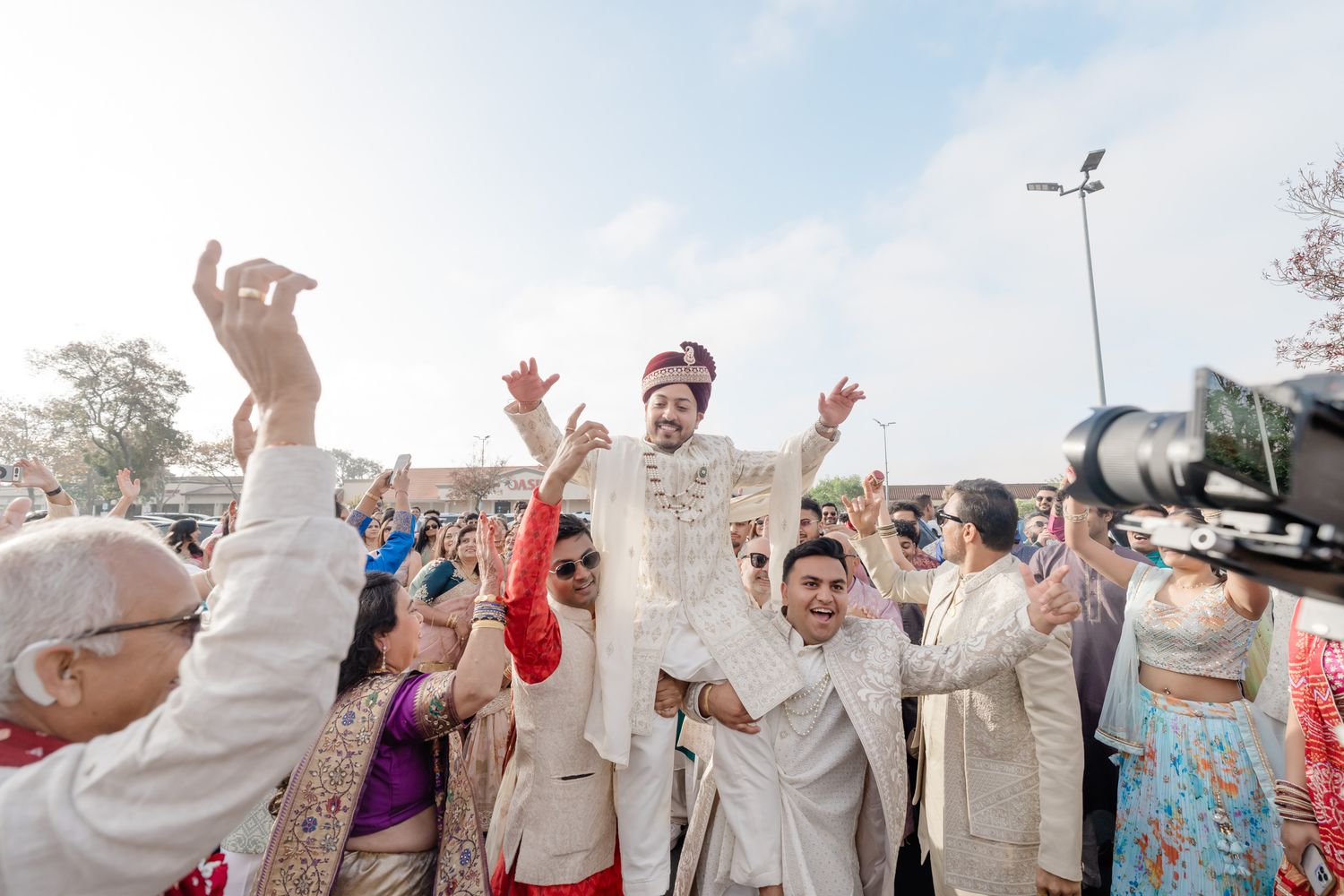 Joyful wedding celebration with guests dancing and cheering during a traditional Indian baraat ceremony outdoors.