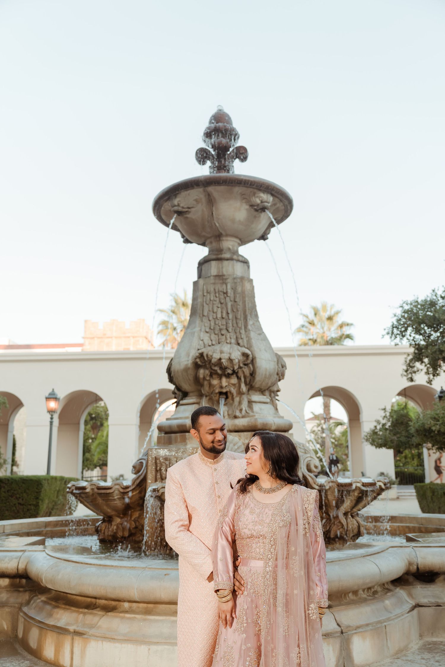 A couple in coordinated pink traditional outfits pose together by an ornate stone fountain in a courtyard.