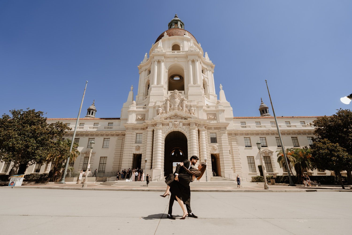 Couple embracing in front of Pasadena City Hall, a grand Beaux-Arts building under a clear blue sky.