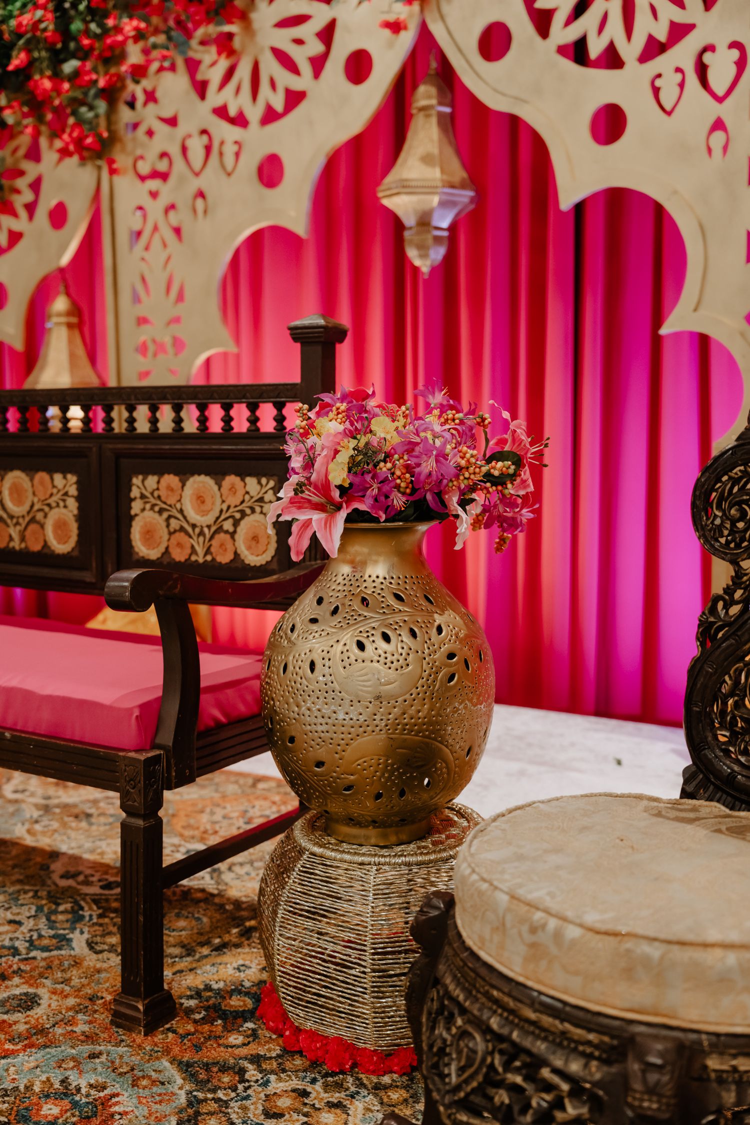 Traditional Indian wedding decor with carved wooden bench, bronze vase and pink floral backdrop with lantern.