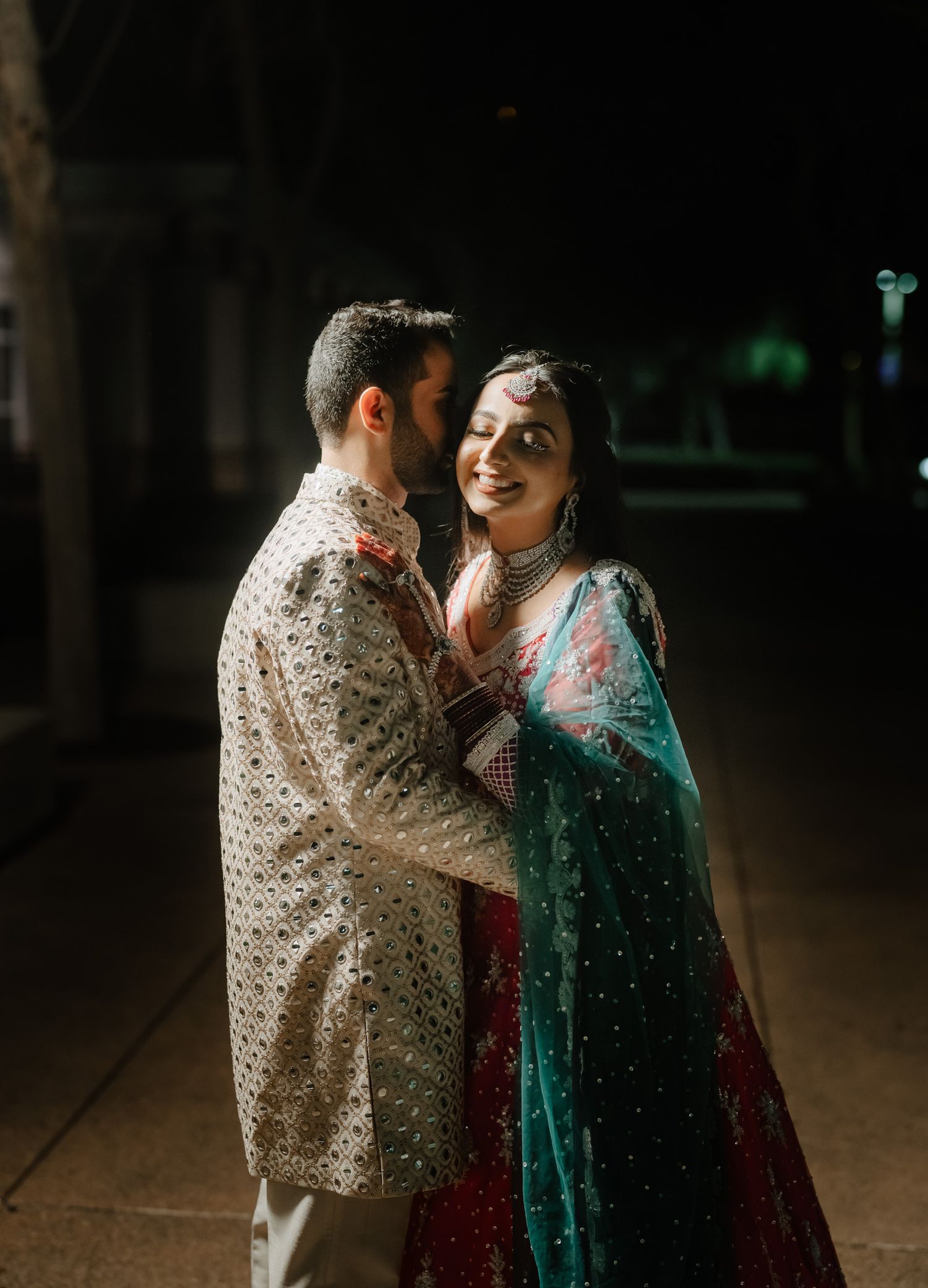 A couple in traditional Indian wedding outfits embraces while sharing a joyful moment under dramatic lighting.