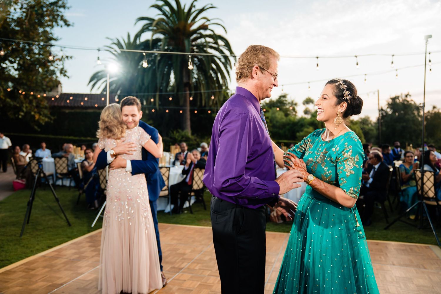 Wedding guests dance together outdoors at dusk during a lively reception celebration.