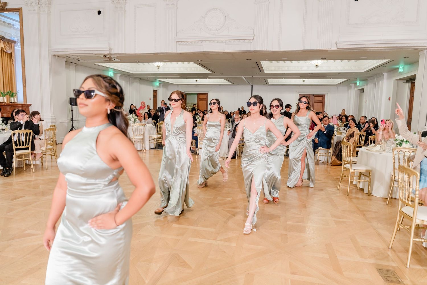 Wedding party members in silver satin dresses performing a choreographed dance routine in a bright reception hall.