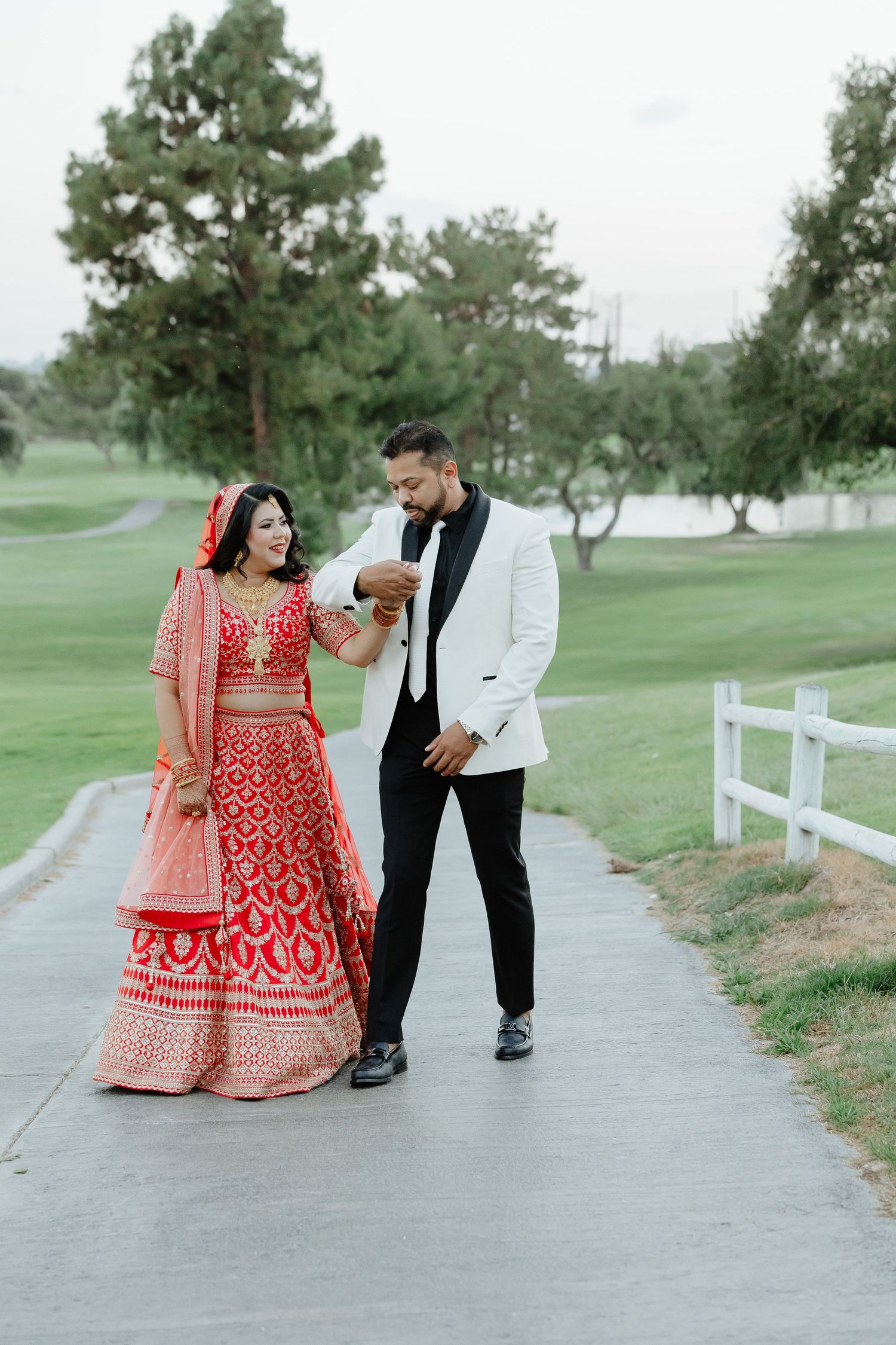 Indian bride in red lehenga and groom in white tuxedo walking together on a path at an outdoor wedding venue.