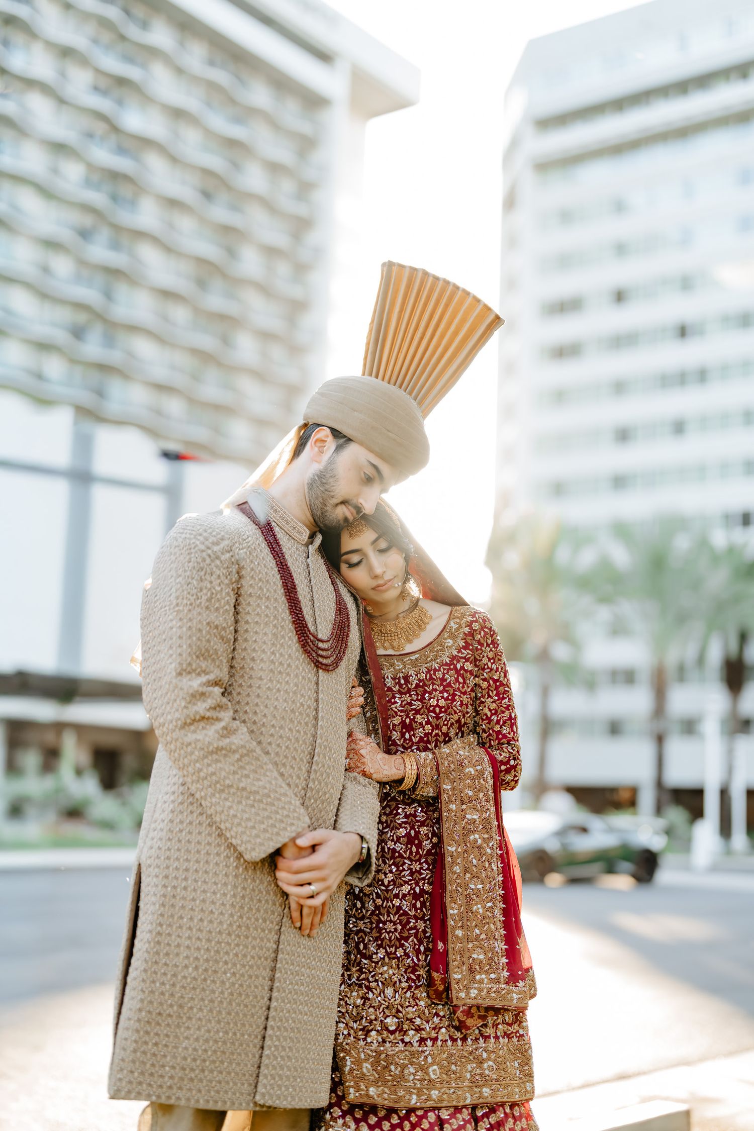 A couple in traditional South Asian wedding attire embrace outdoors near modern architecture.