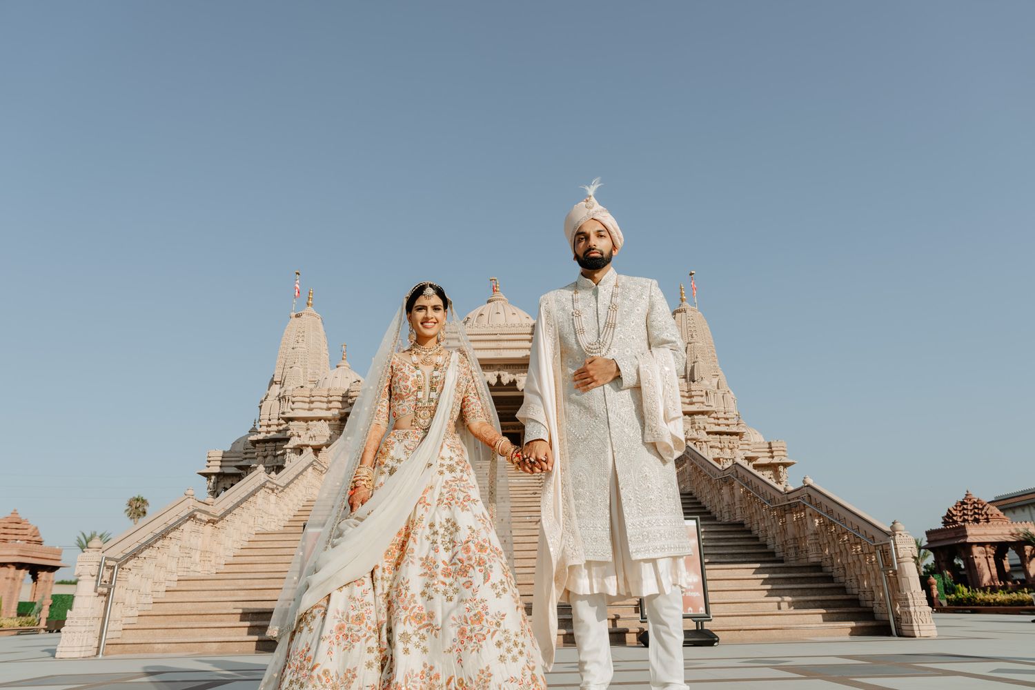 A couple in traditional Indian wedding attire standing in front of an ornate temple architecture.