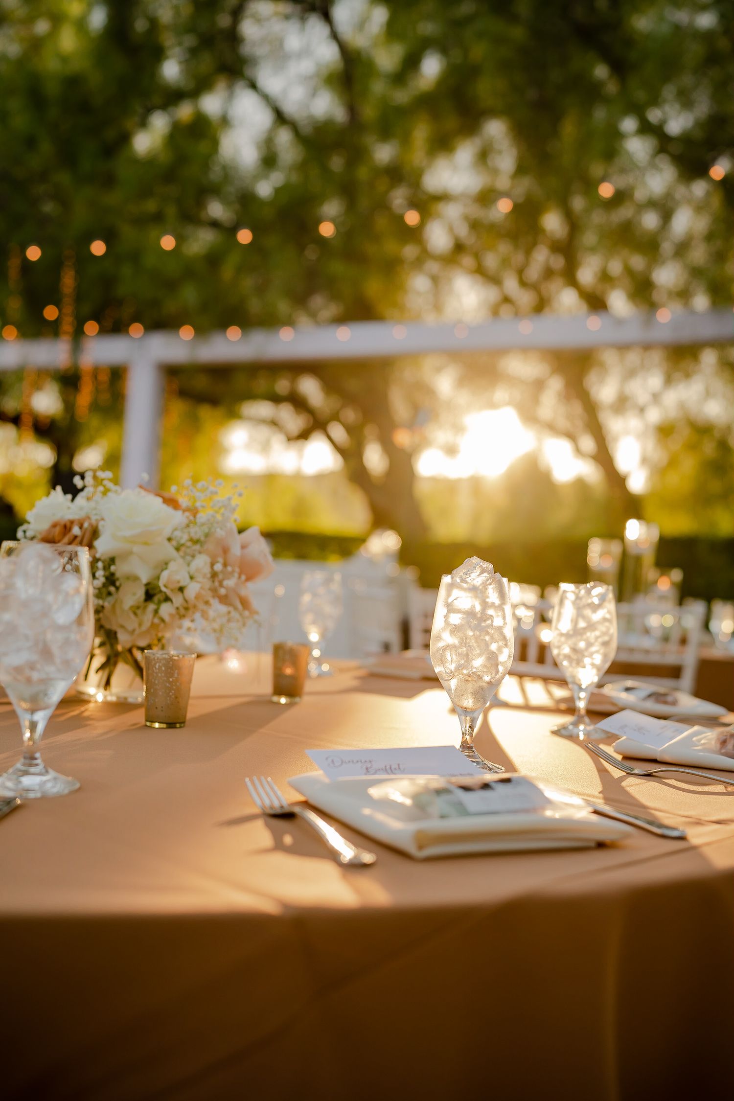 Romantic outdoor wedding table setting at sunset with glowing string lights and floral centerpieces.