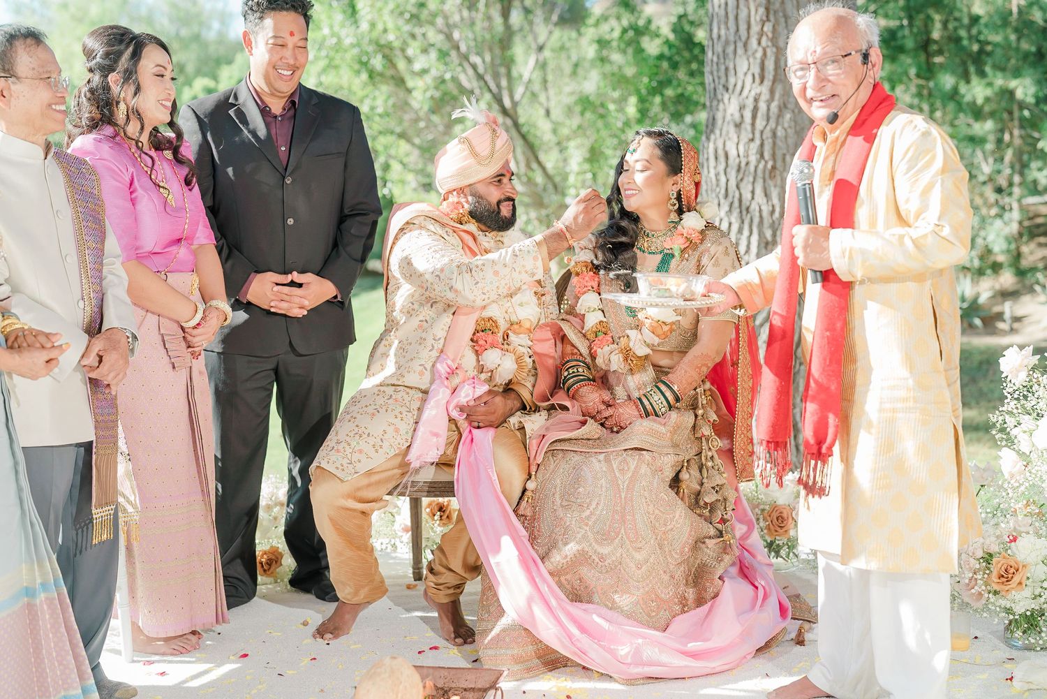 A traditional wedding ceremony takes place outdoors with attendees wearing formal attire in pink and neutral tones.