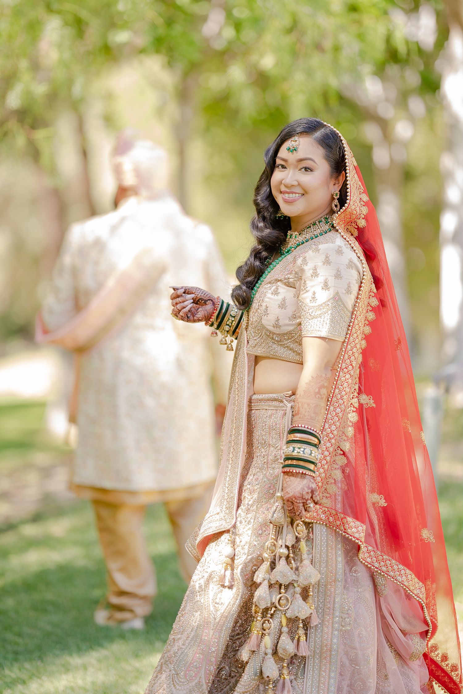 A couple in elegant Indian wedding attire poses together on a sunny day in a lush garden setting.