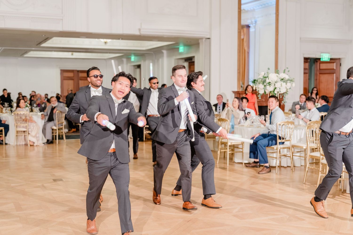 Groomsmen in gray suits perform a synchronized dance move during a wedding reception celebration.