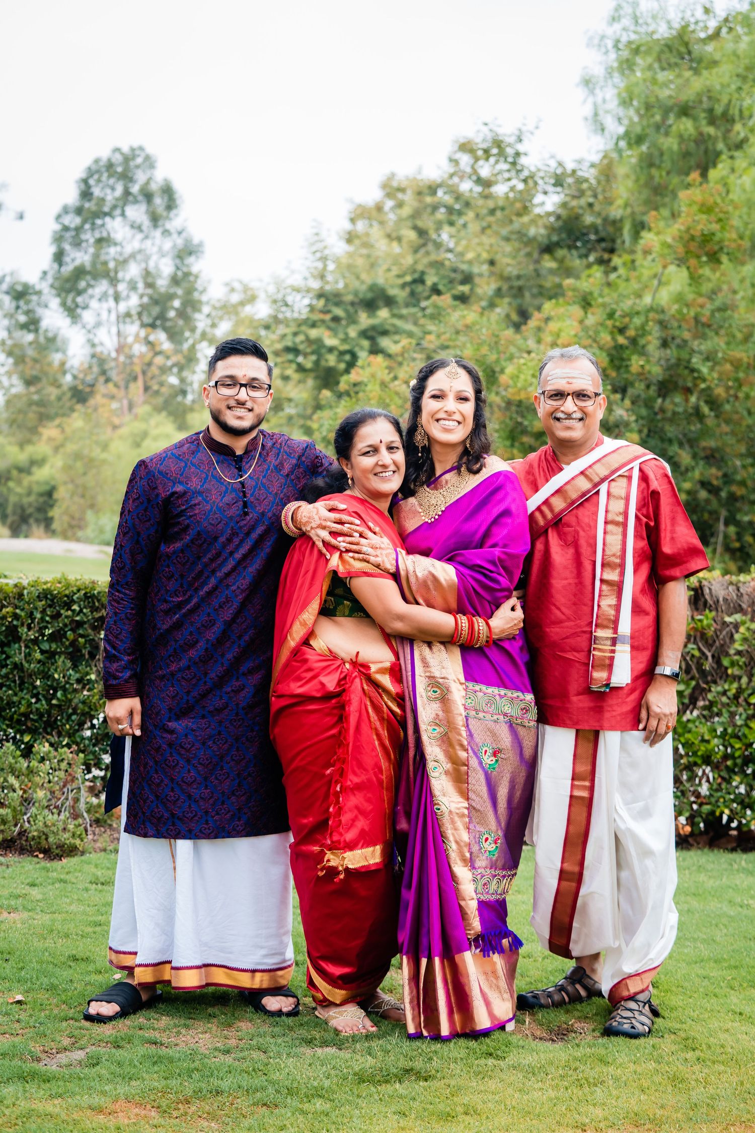 A family group poses outdoors on grass wearing traditional South Indian attire in purple, red and white colors.