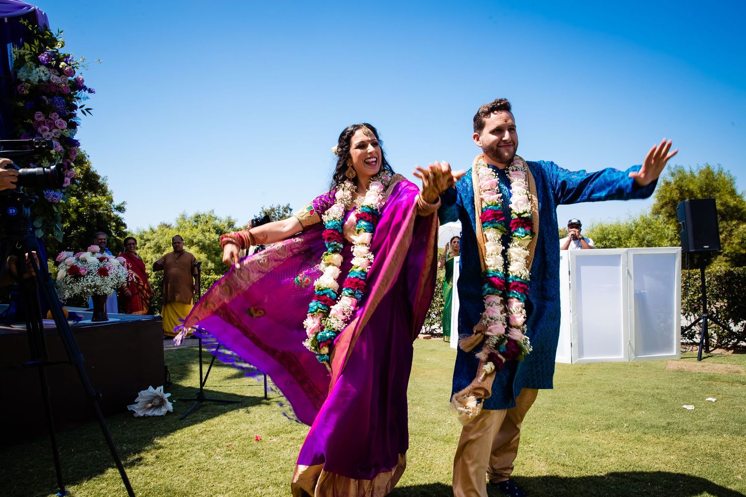 Wedding party poses playfully on a sunny lawn with flowing purple dresses creating dynamic movement.