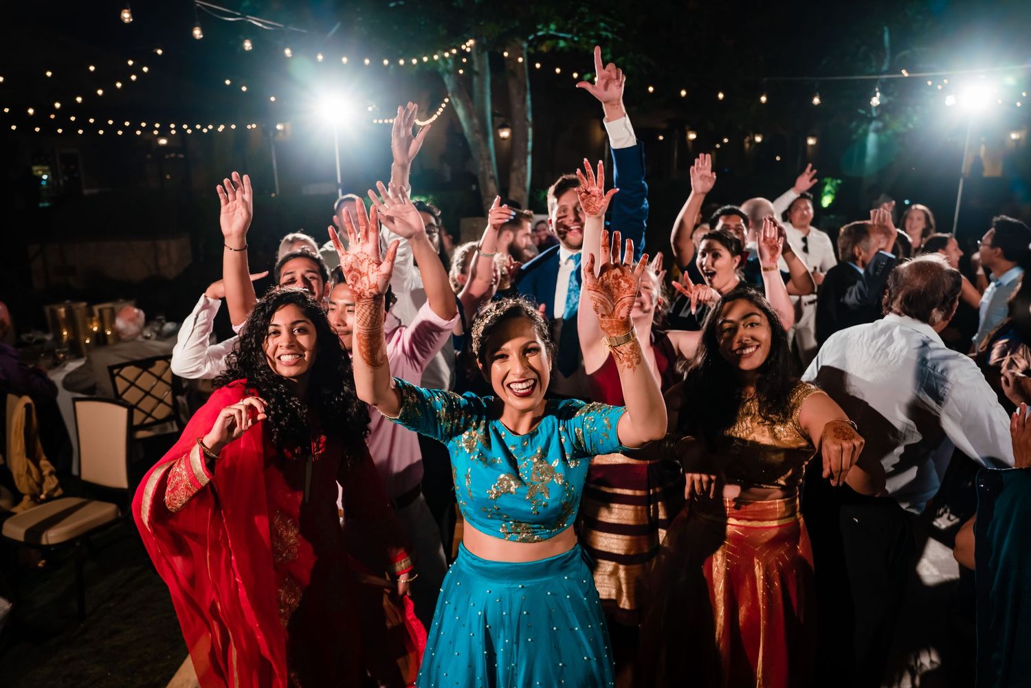 A lively outdoor party scene at night with string lights overhead as a crowd dances and celebrates with raised hands.