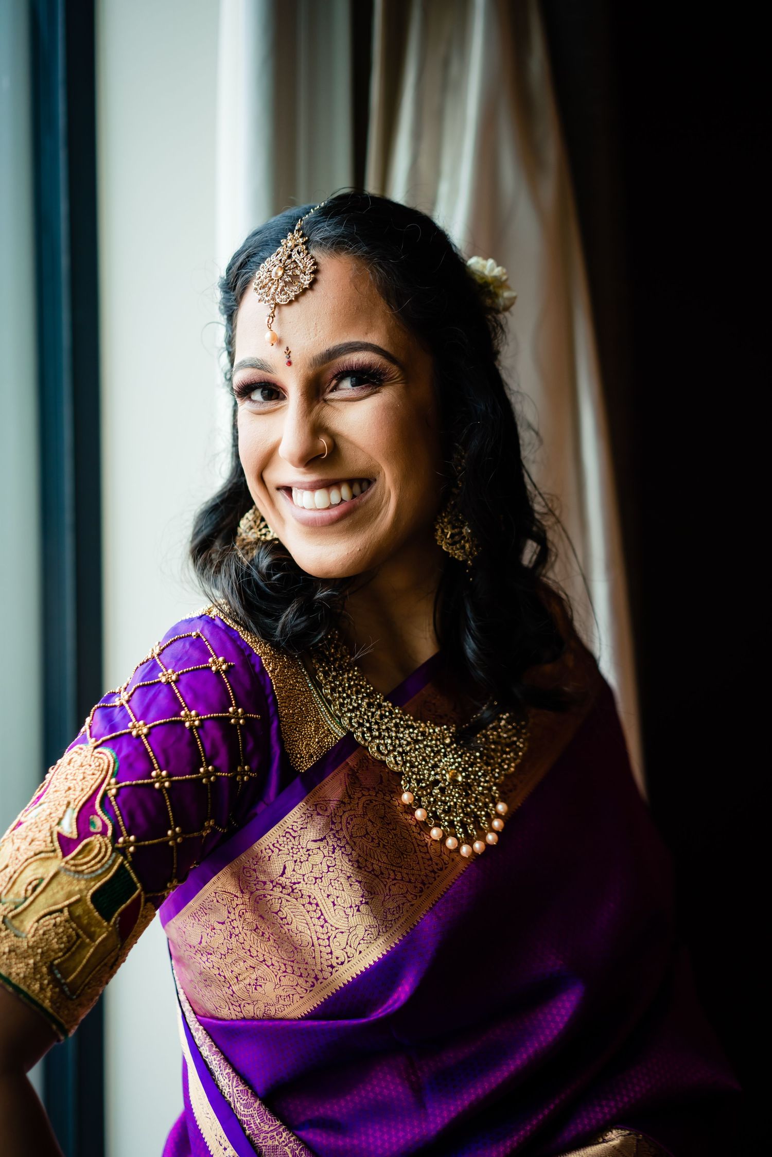 Traditional Indian bride in deep purple and gold silk saree with ornate jewelry smiles during wedding preparations.