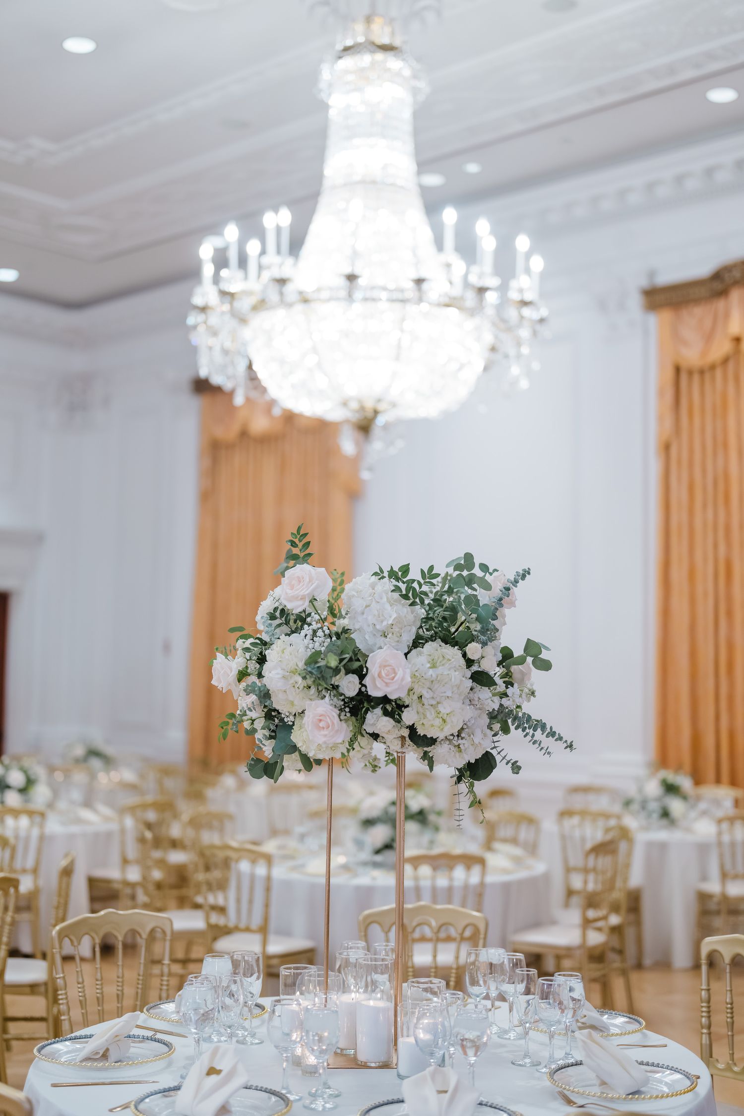 Elegant wedding reception tables with crystal chandeliers, gold chairs and white floral centerpieces.