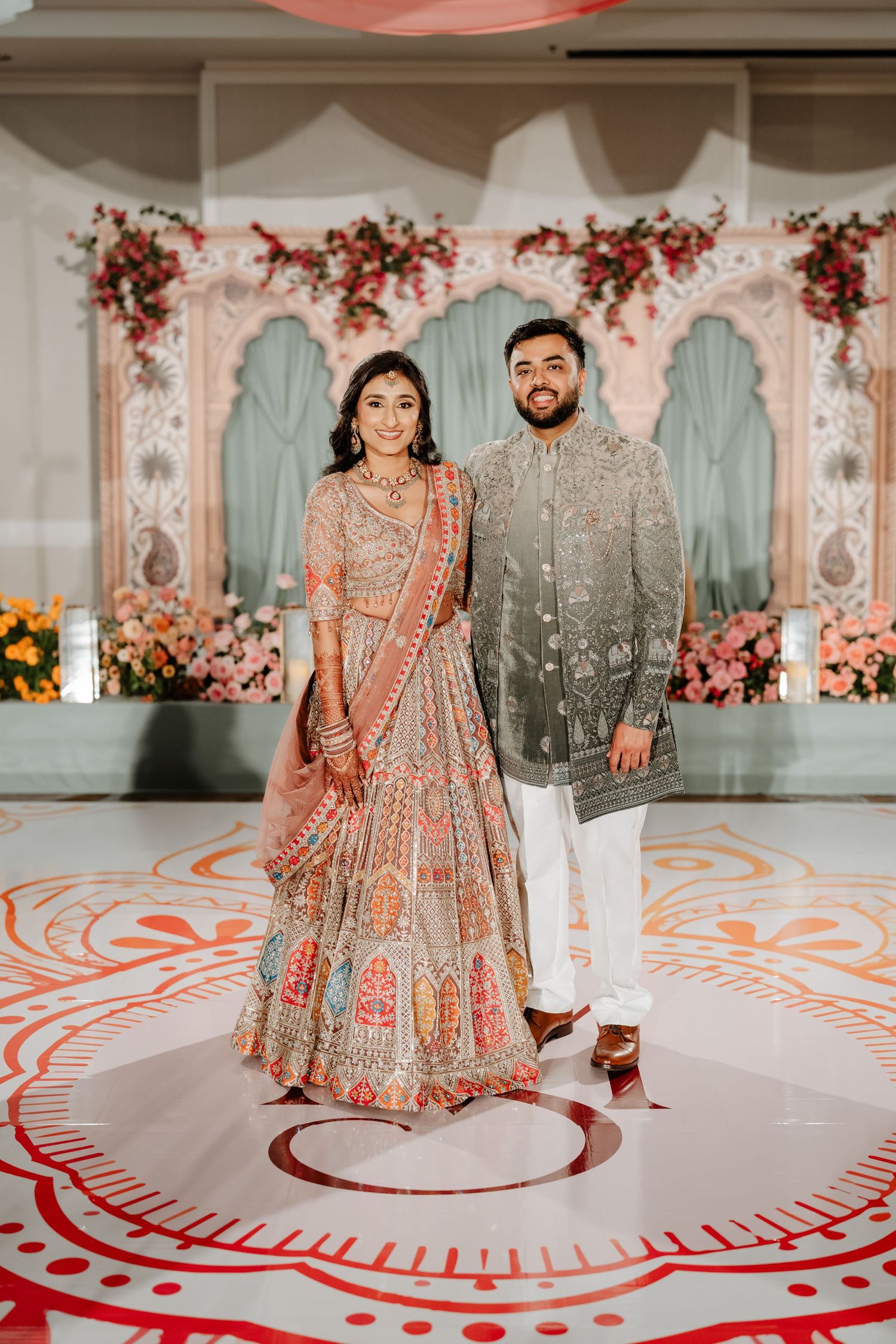 Couple in traditional Indian wedding attire posing on decorated dance floor with floral backdrop.