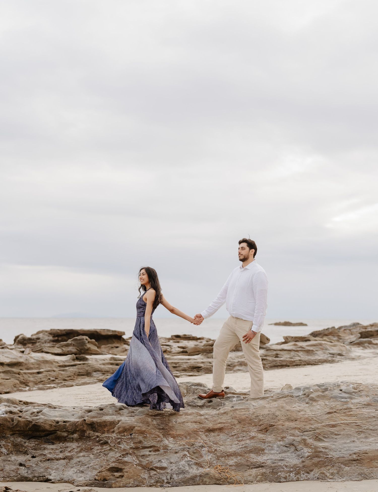 Couple holding hands walking on rocky beach, woman in flowing blue dress, man in white shirt and khaki pants.