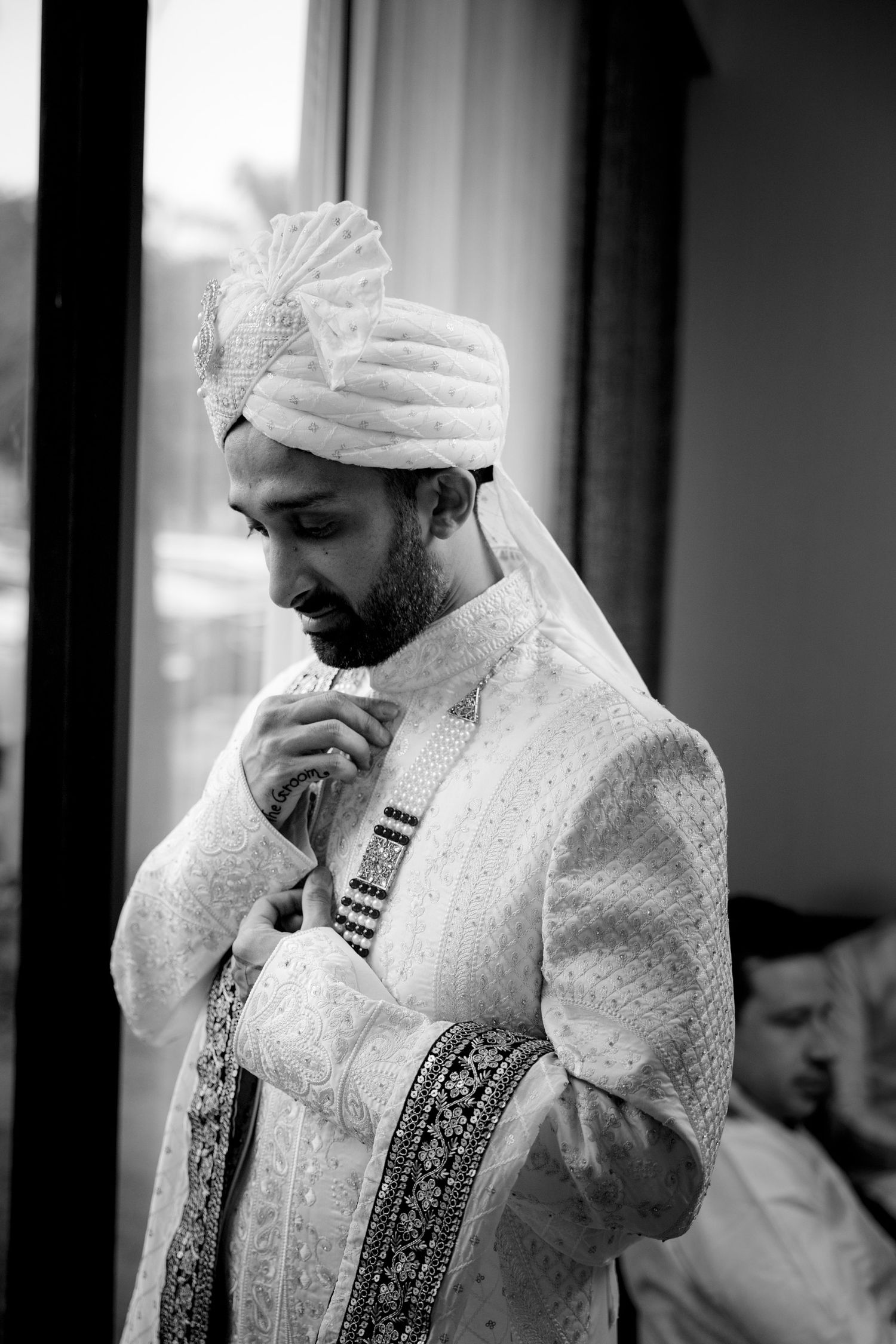 Black and white photo of an Indian groom in white sherwani and turban adjusting his outfit near a window.