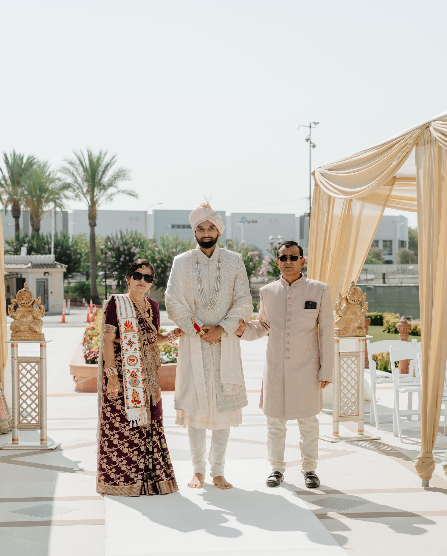 Wedding guests stand under elegant cream-colored canopies at an outdoor venue with palm trees in the background.