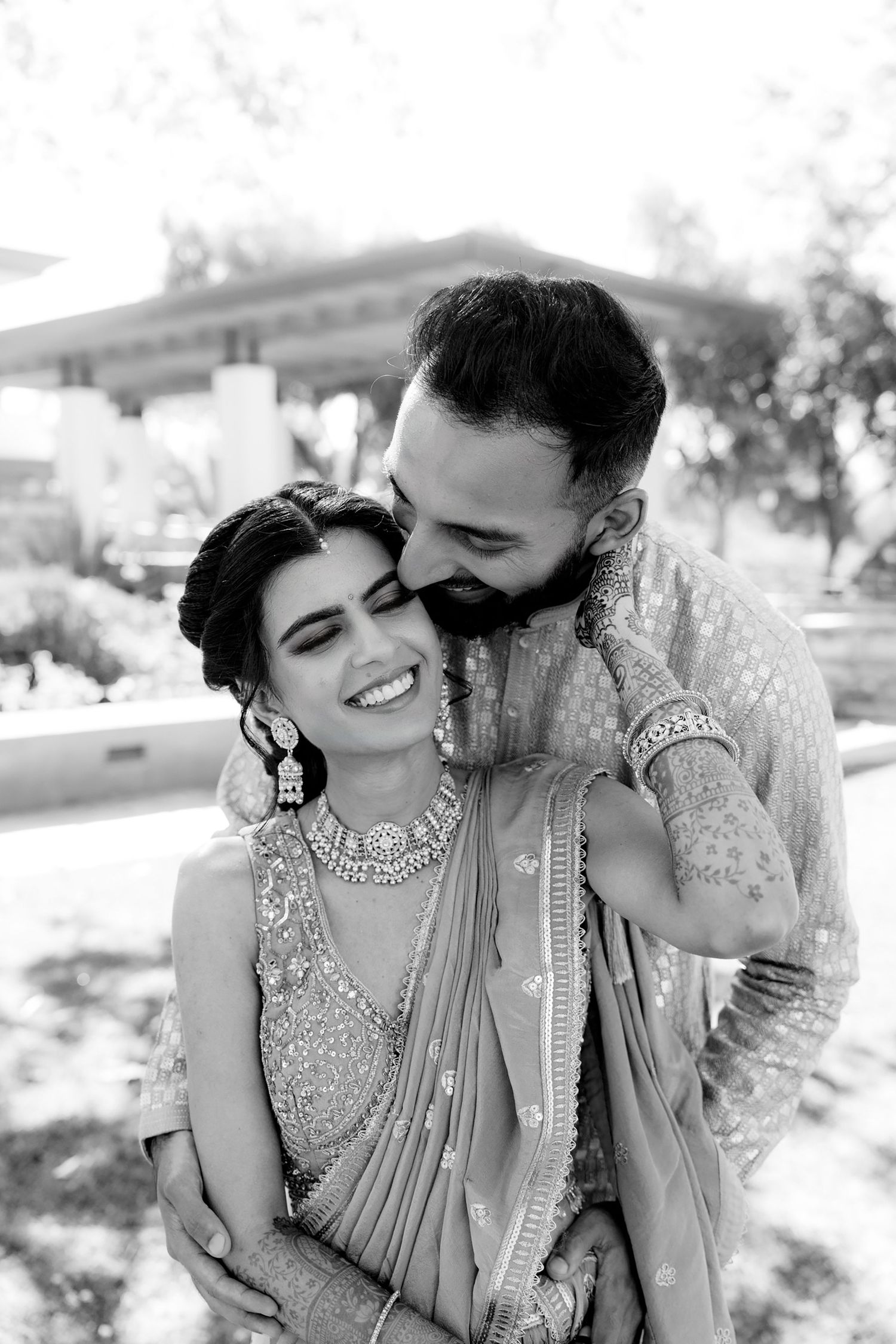 A romantic black and white photo of a couple in traditional Indian wedding attire sharing an intimate moment outdoors.