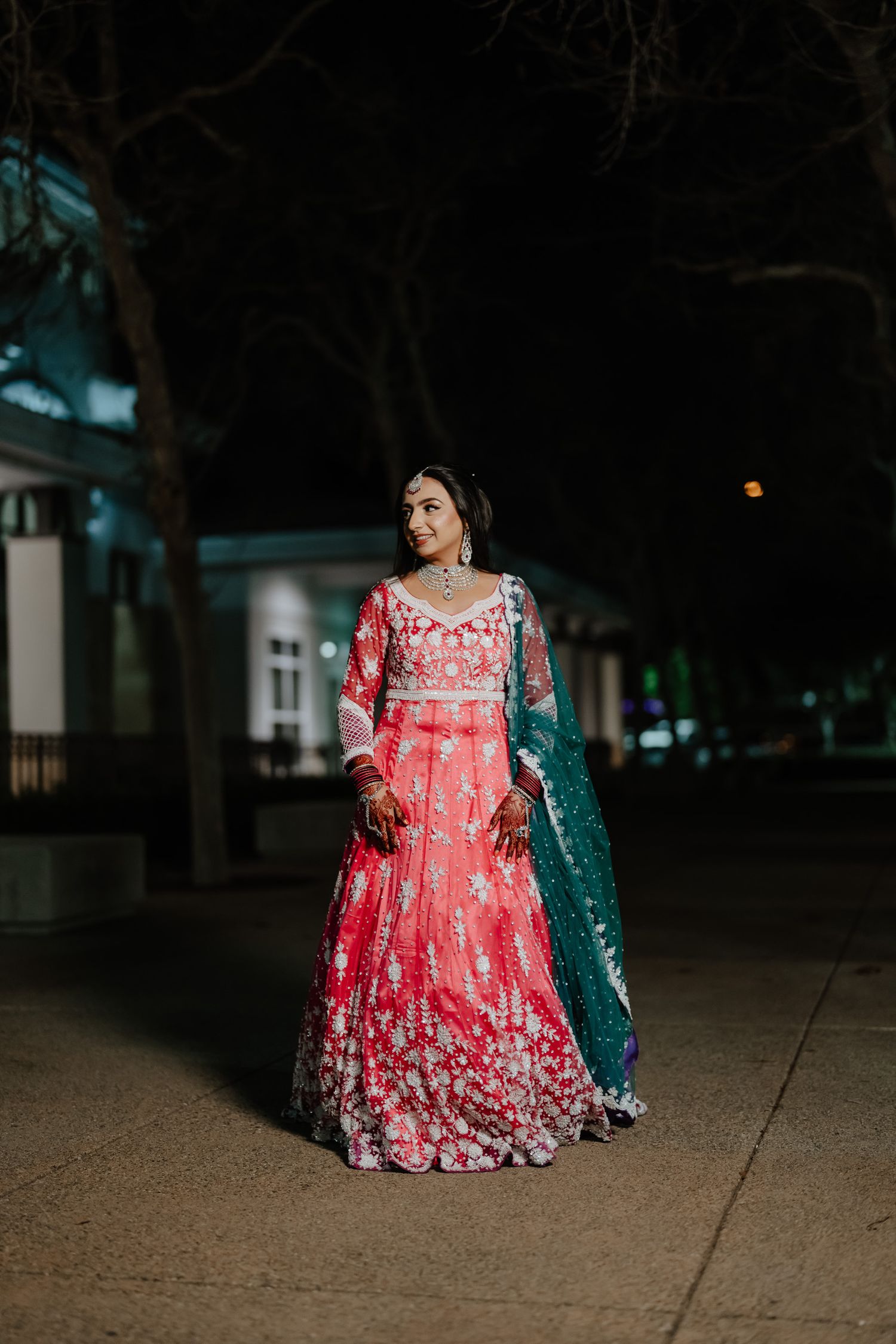 A woman stands outdoors at night wearing a stunning red and white embroidered lehenga with a teal dupatta.