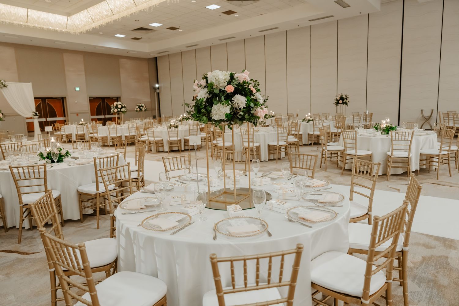 Spacious wedding reception venue with white round tables and gold chiavari chairs arranged for celebration.
