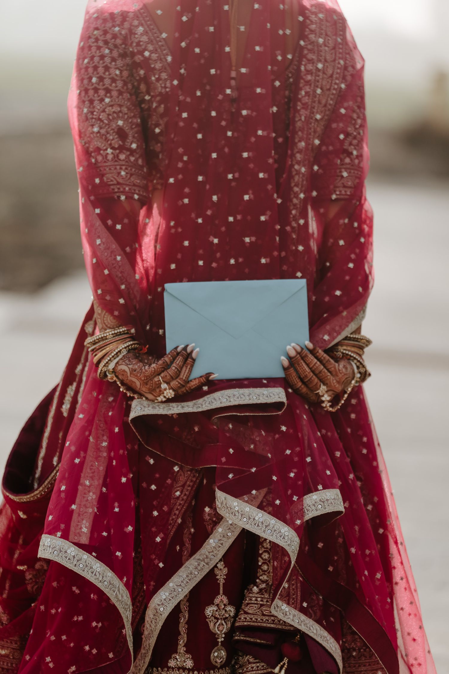 Traditional Indian bride in red embroidered dress holding a light blue envelope with henna decorated hands.