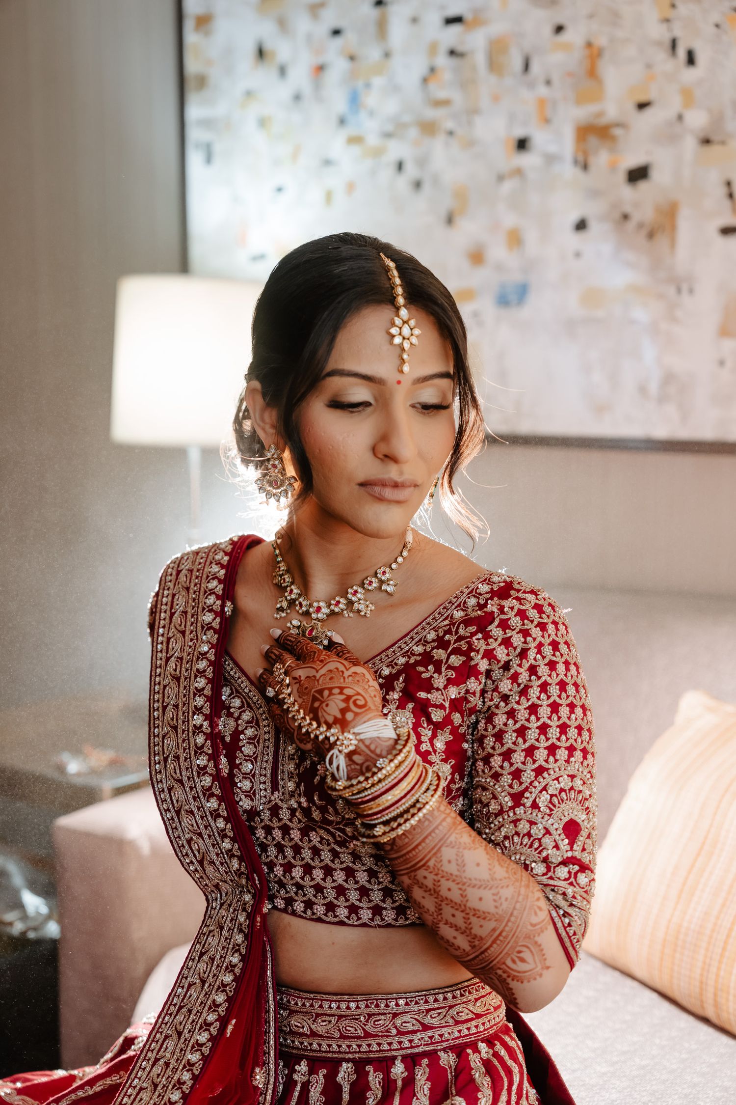 A traditional Indian bride in a red embroidered lehenga and ornate jewelry poses elegantly in a softly lit room.