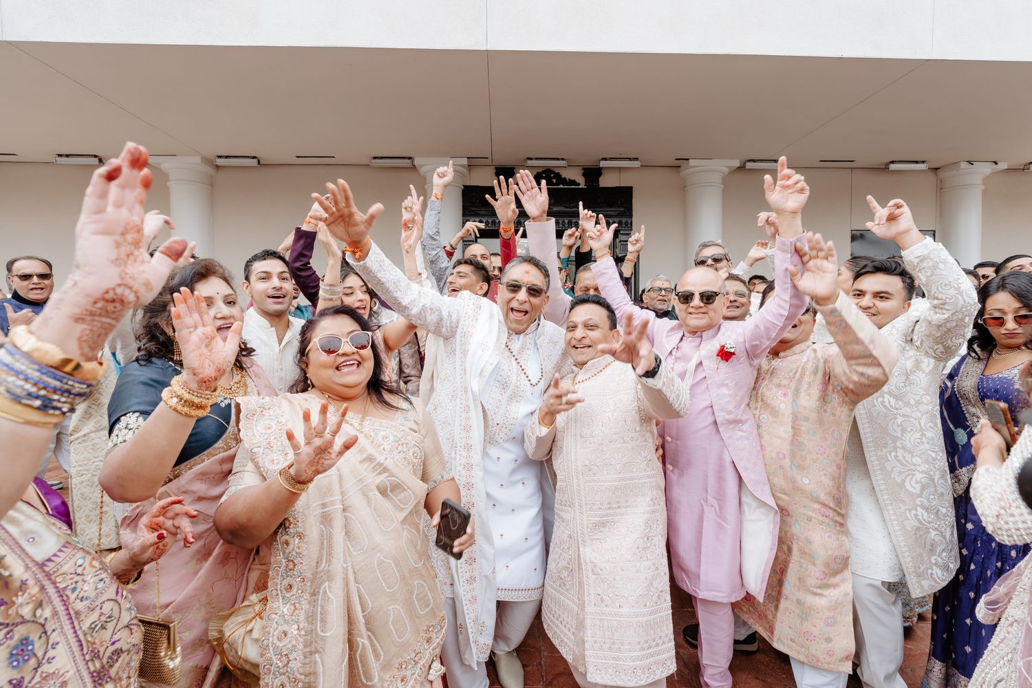 Large group of wedding guests in white attire raise their hands in celebration during a joyous reception moment.