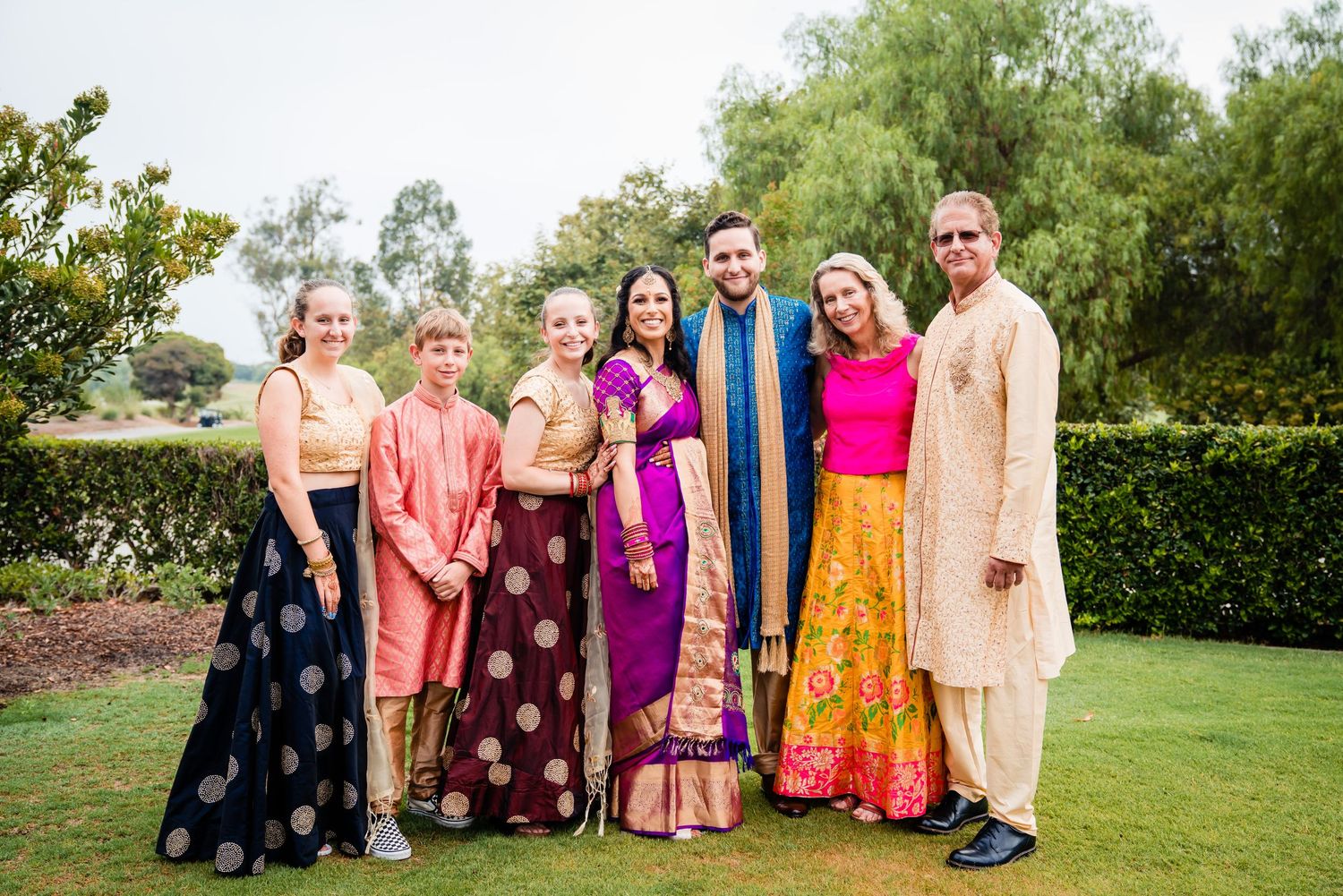A group in colorful formal attire stands together on a green lawn for a special occasion photo.