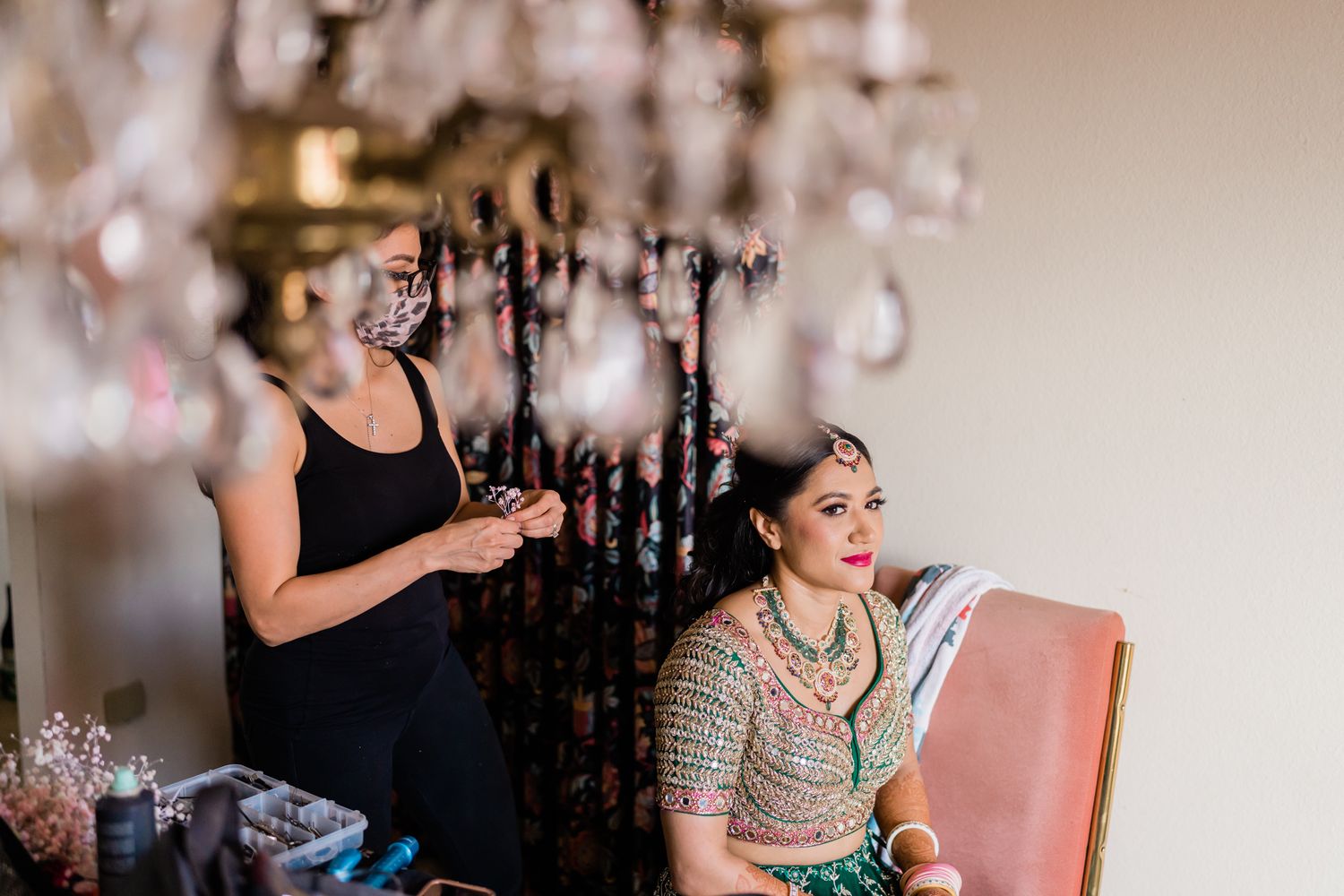 A stylist works on a bride's hair while she sits in a pink chair under an elegant chandelier.