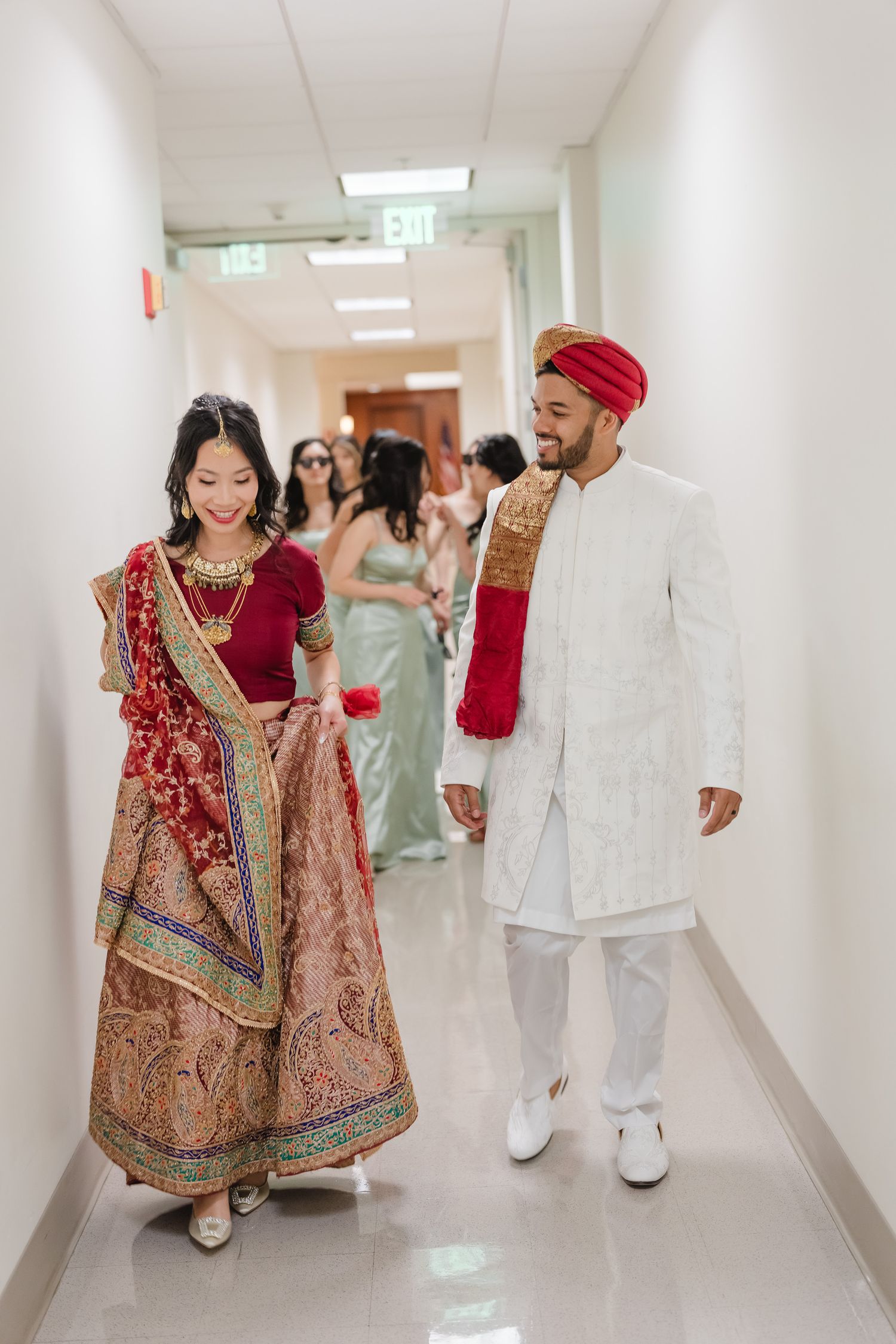A bride in traditional red and gold South Asian wedding attire walks joyfully down a bright hallway.